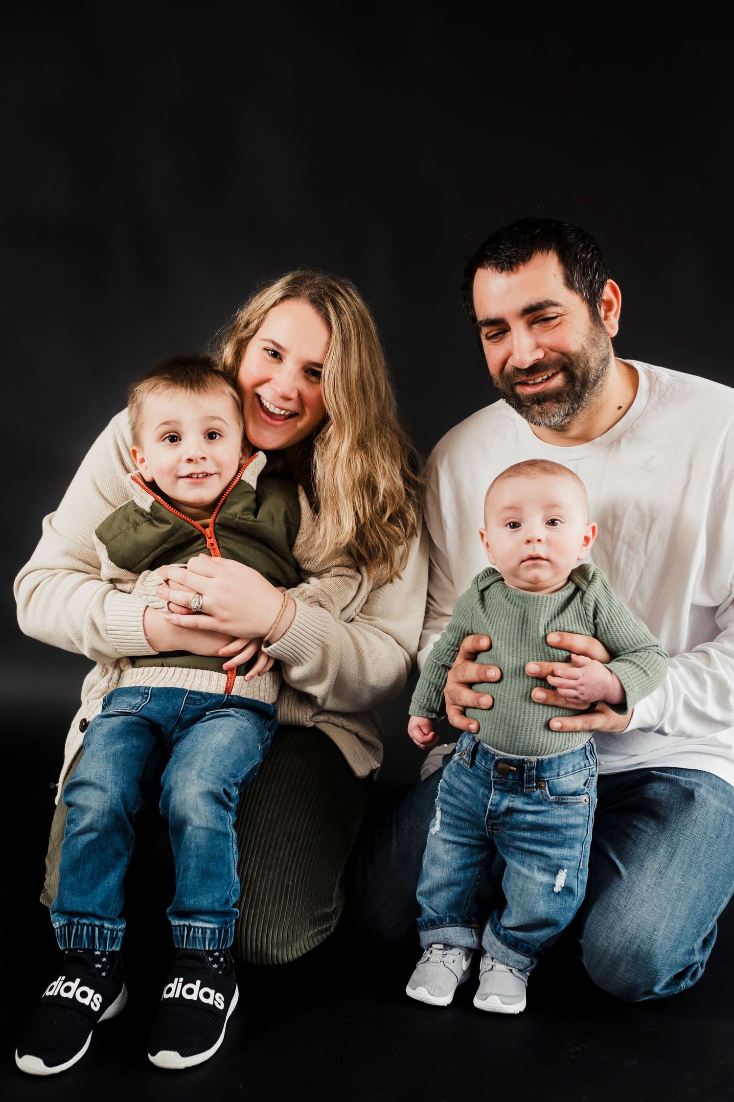 Family portrait with parents and two young children wearing coordinated casual outfits against a dark background.