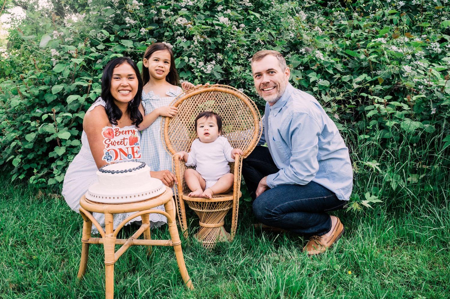 A family poses together outdoors on wicker chairs surrounded by green foliage on a summer day.