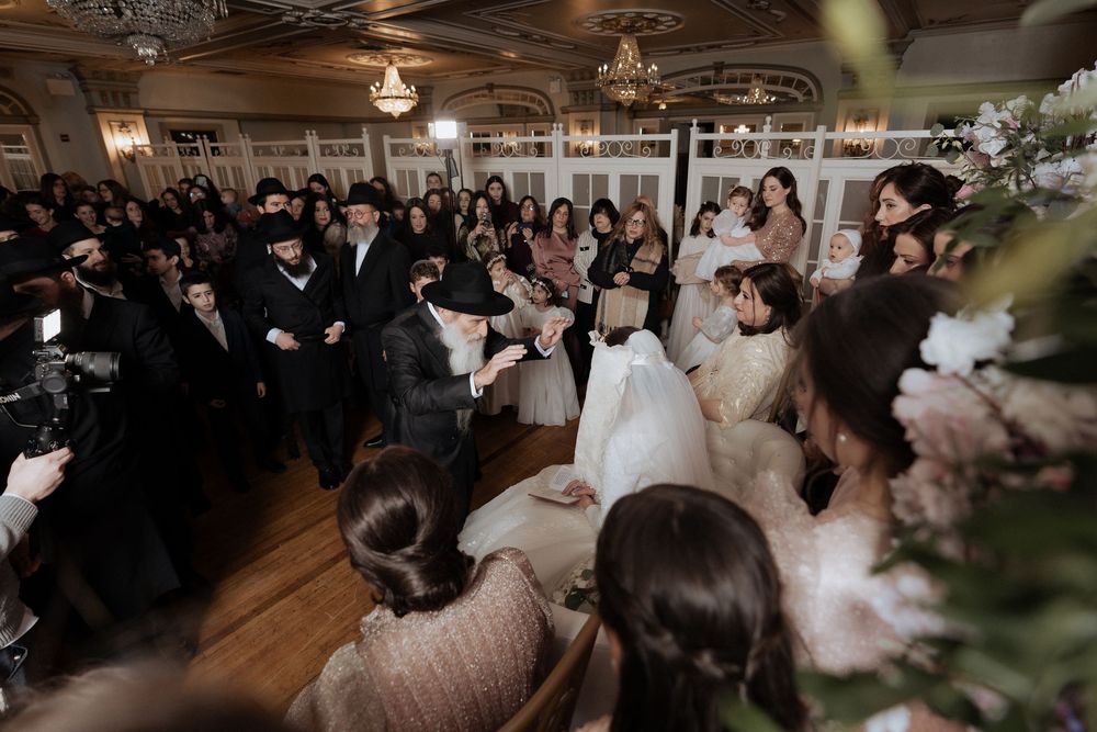 Traditional Jewish wedding celebration showing guests dancing in a crowded ballroom with elegant chandeliers.