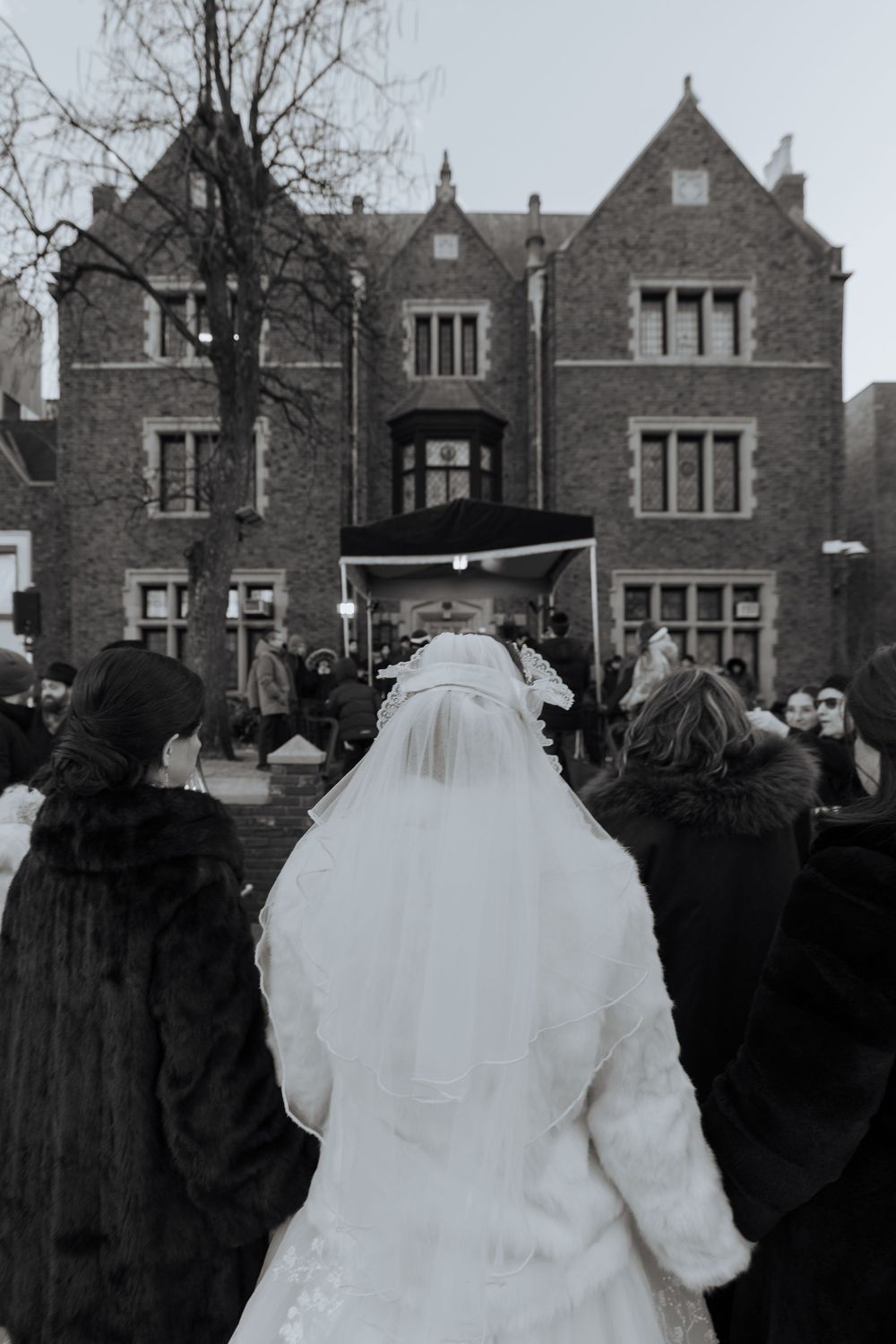 Bride in white coat and veil approaches Gothic-style building surrounded by winter trees.