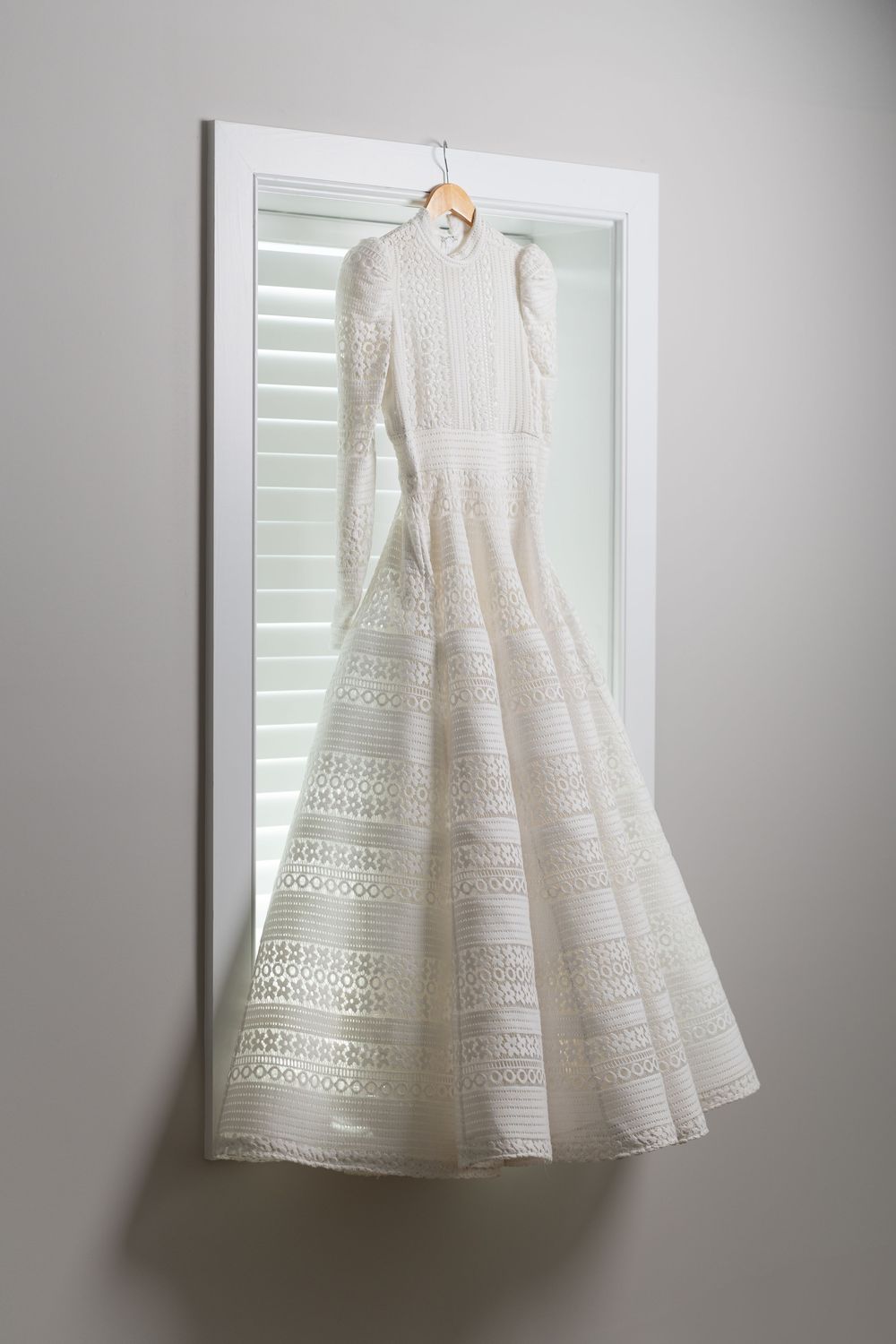 An elegant white lace wedding dress with intricate patterns hanging on display against a window with shutters.