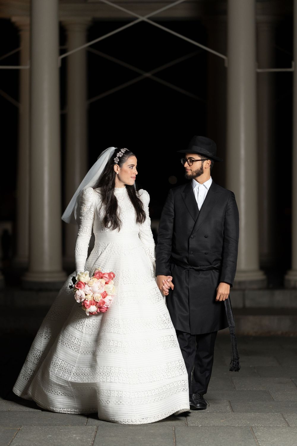 A bride in a detailed lace gown and groom in formal attire stand between classic columns at night.