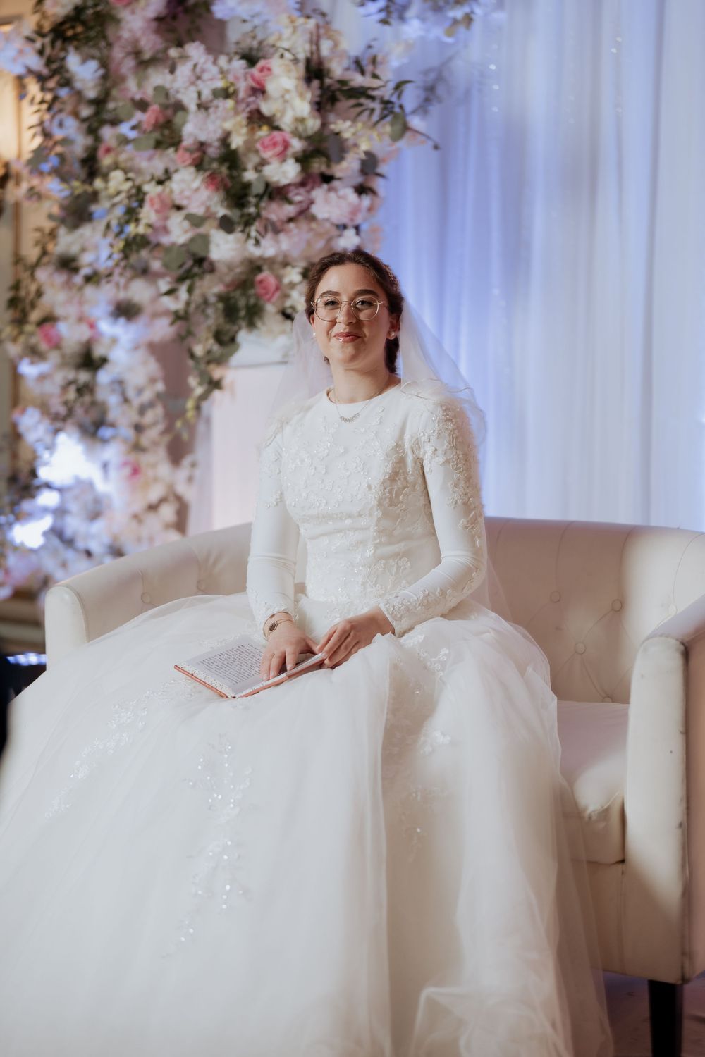 Bride in long-sleeved lace wedding gown sits elegantly on cream couch with floral backdrop.