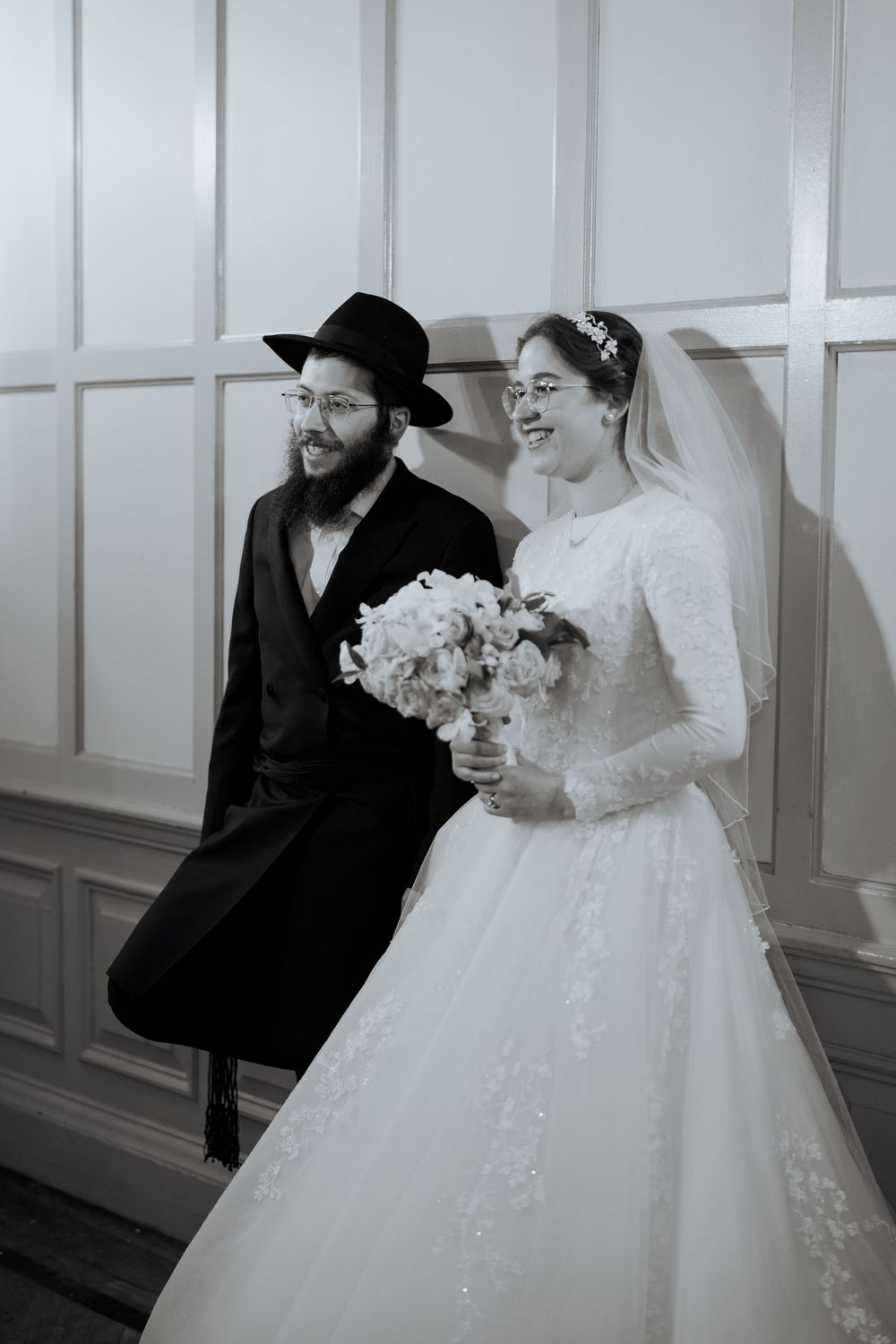 Orthodox Jewish bride and groom pose together, she holding a white bouquet in elegant wedding attire.