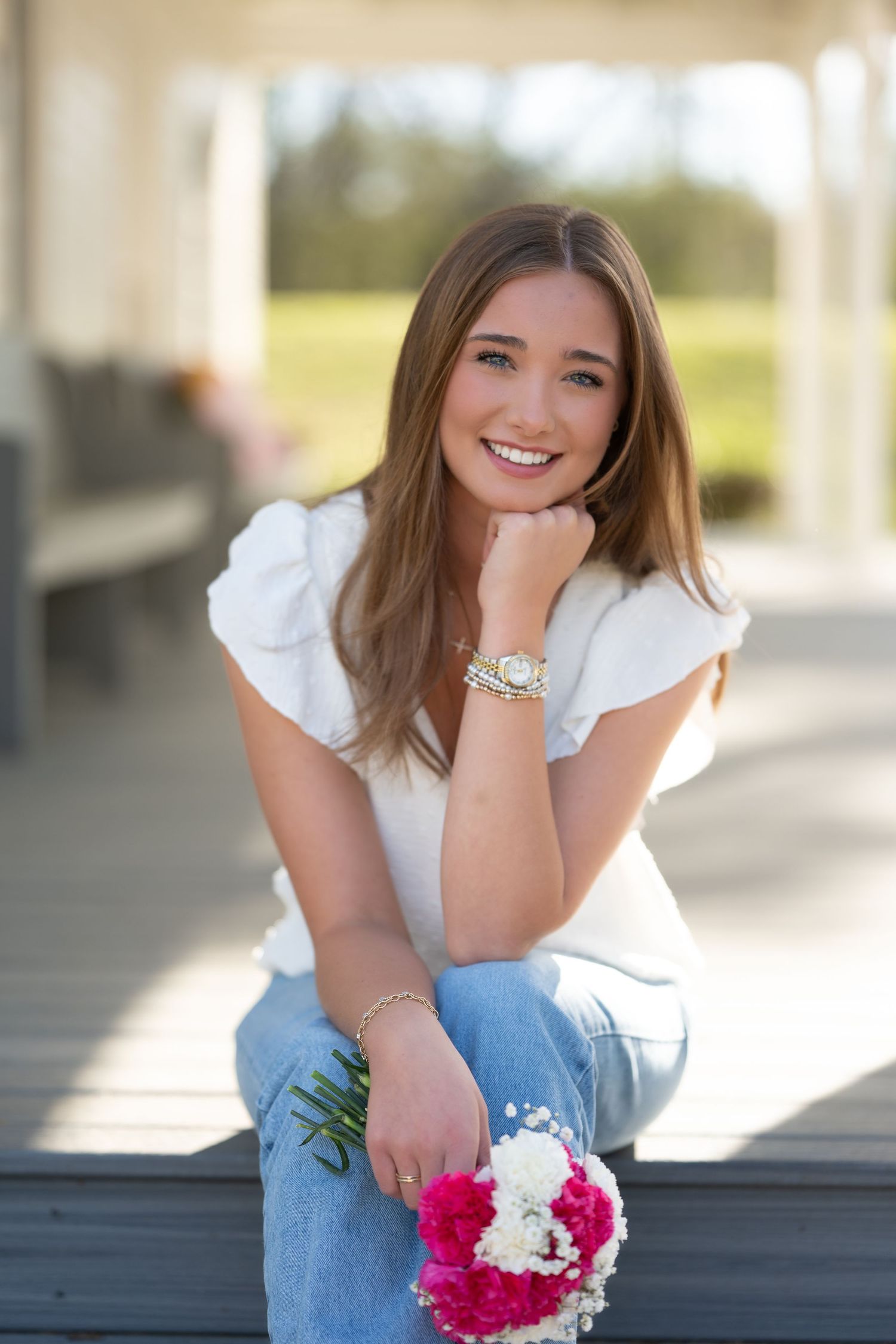 Young woman in white blouse and jeans sits on porch steps holding pink and white flowers while smiling.