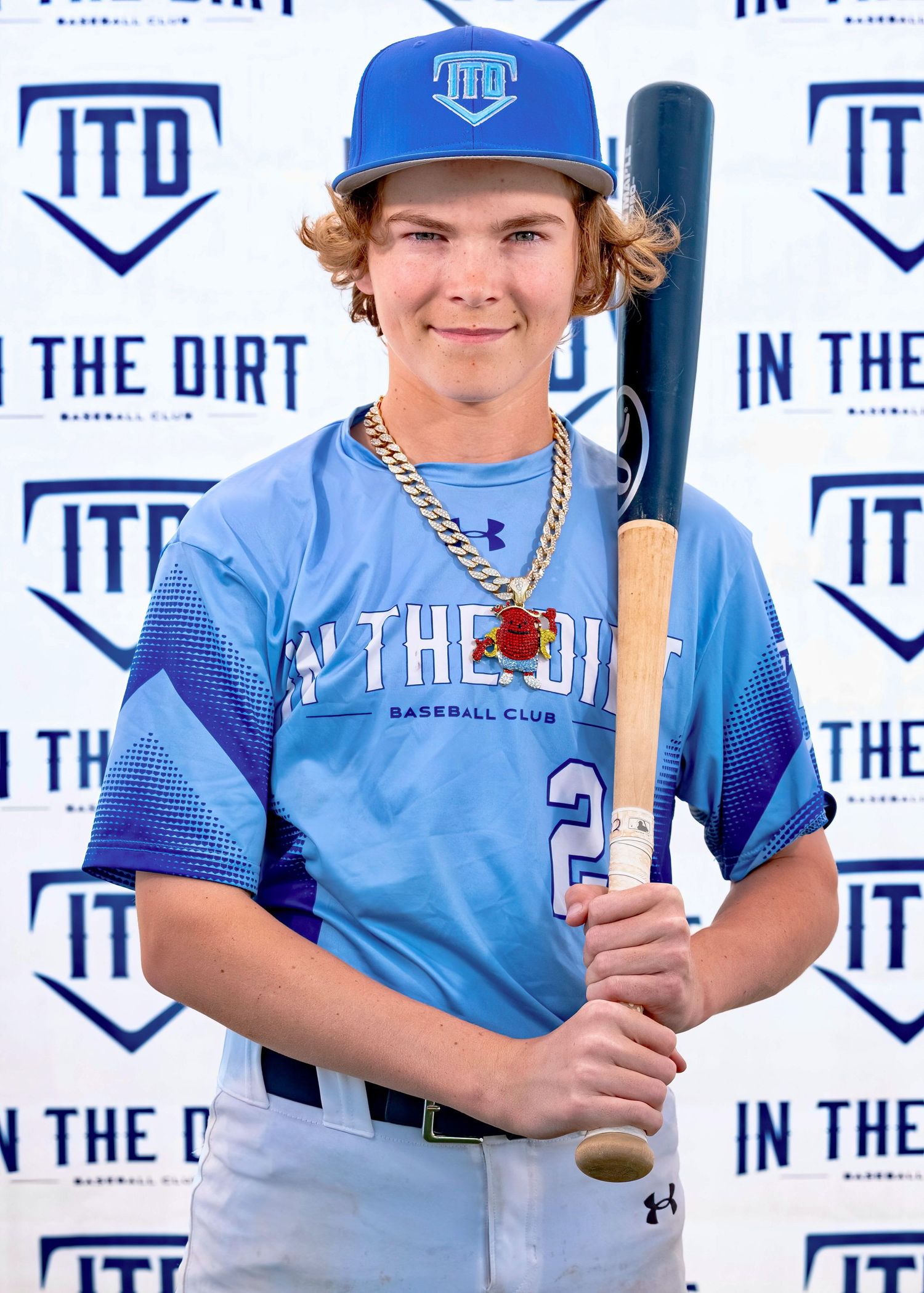Baseball player in light blue jersey and blue cap poses with bat against ITD branded backdrop.