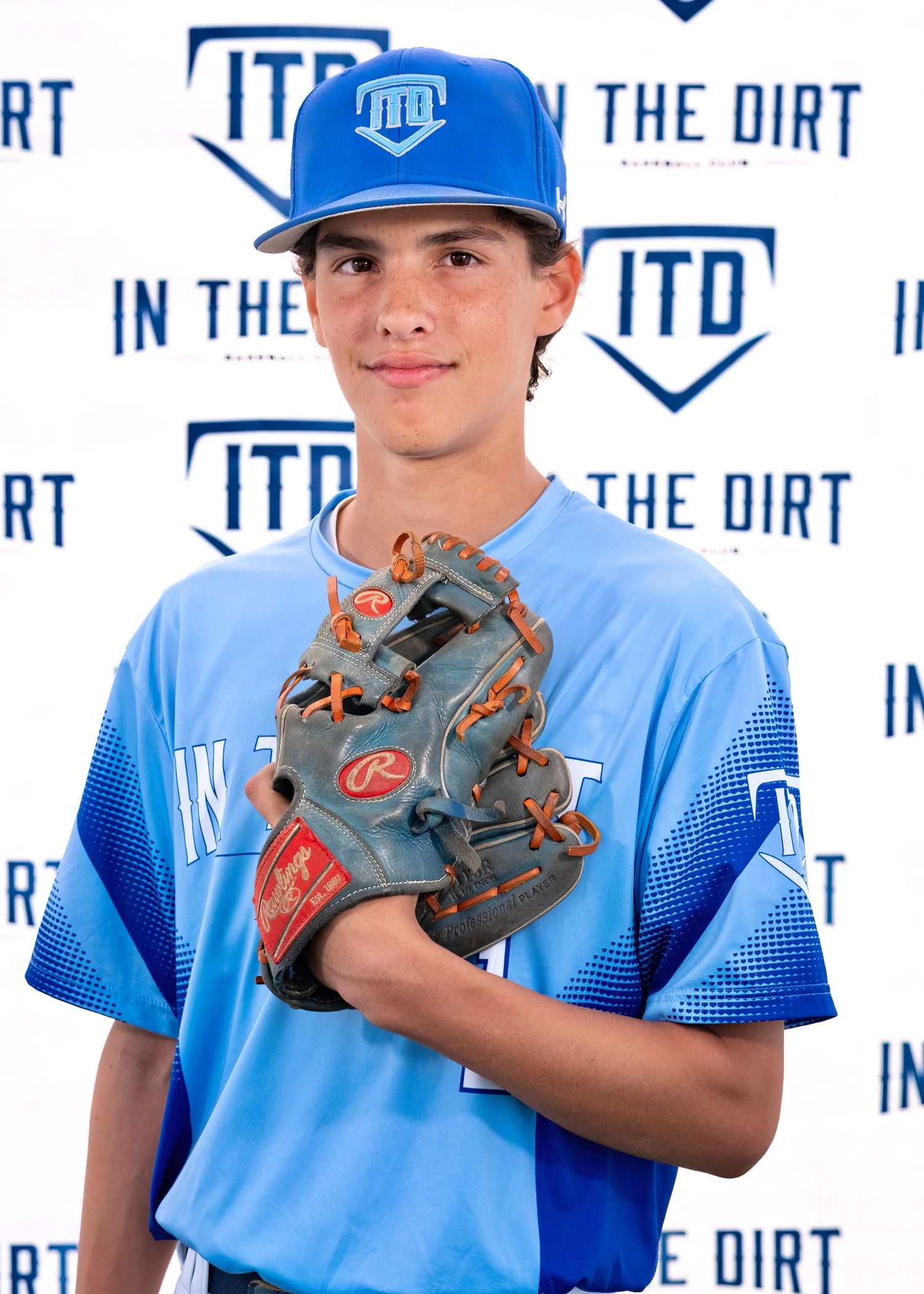 Baseball player wearing light blue uniform and cap with "ITD" logo holds up gray and orange baseball glove against white backdrop.