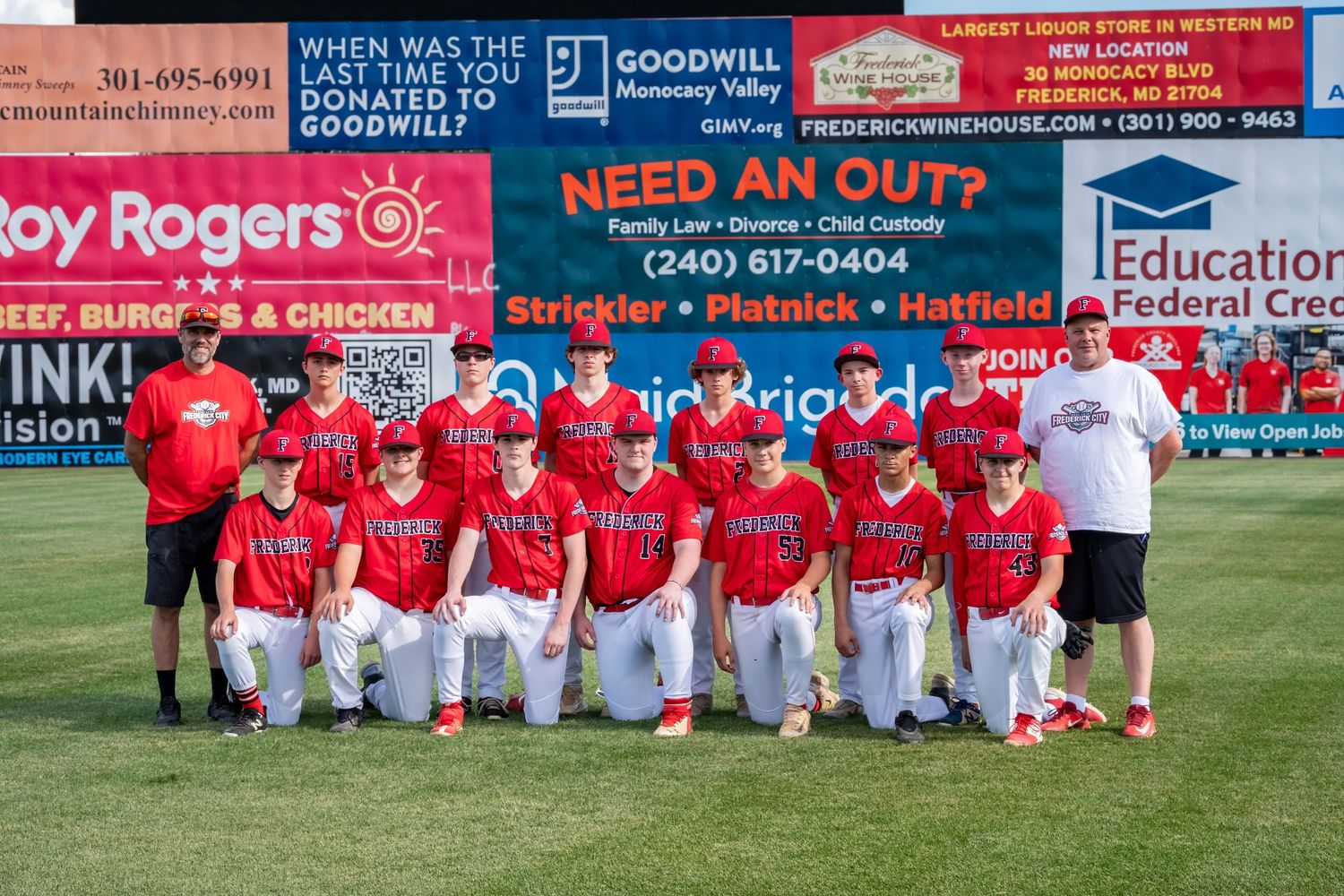 A youth baseball team in red uniforms poses for a group photo on a baseball field with sponsors and banners behind them.