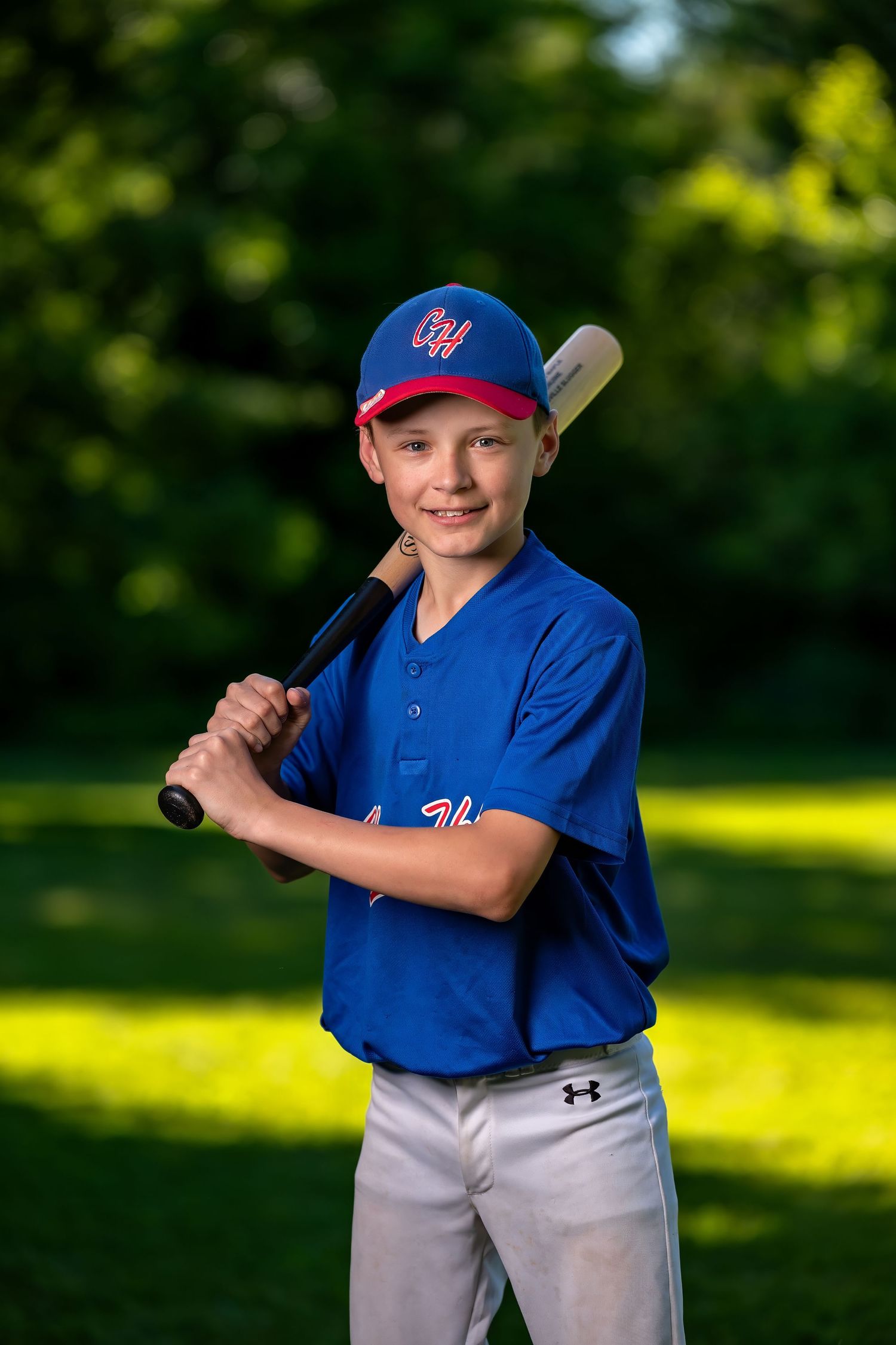 A youth baseball player in a bright blue uniform and cap poses with a bat against a blurred green background.
