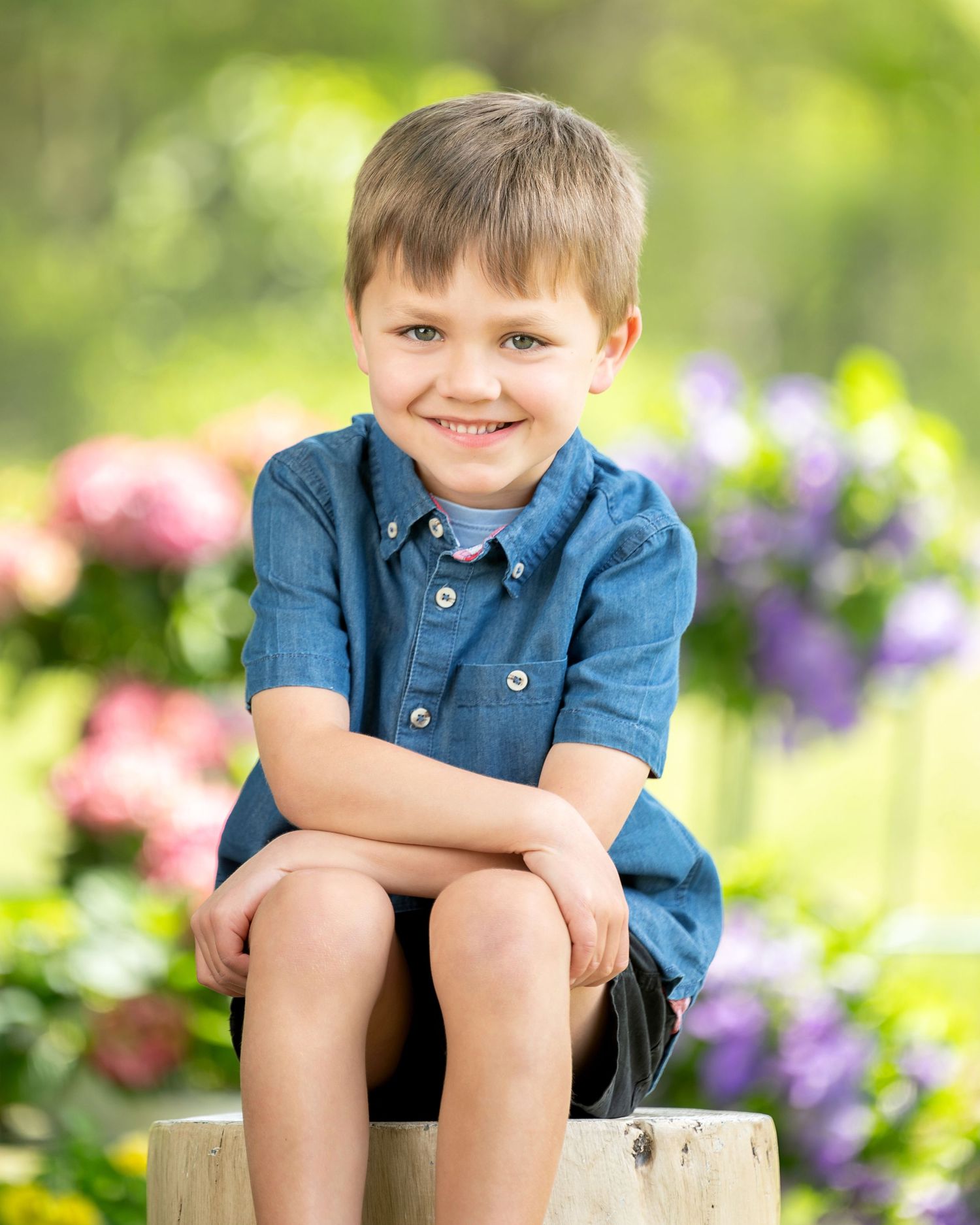 A young child in a striped shirt sits outdoors on a wooden surface surrounded by colorful garden flowers.