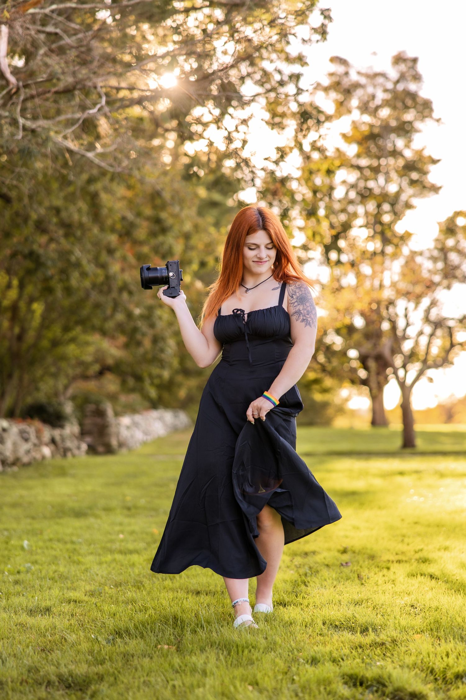 Photographer in black dress poses with camera in sunlit grassy field.