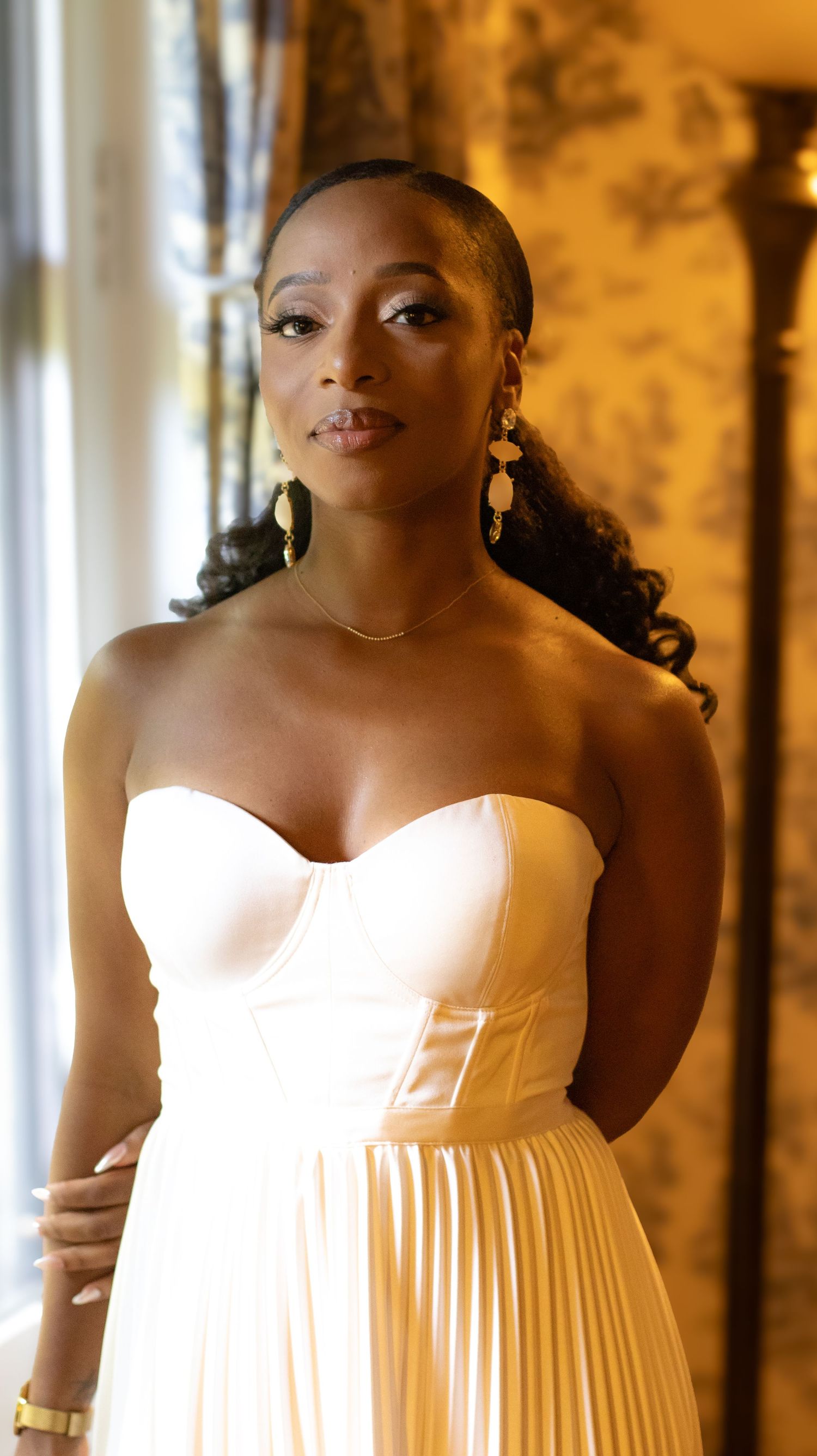 Elegant woman in a white pleated gown with statement earrings and flowing hair.