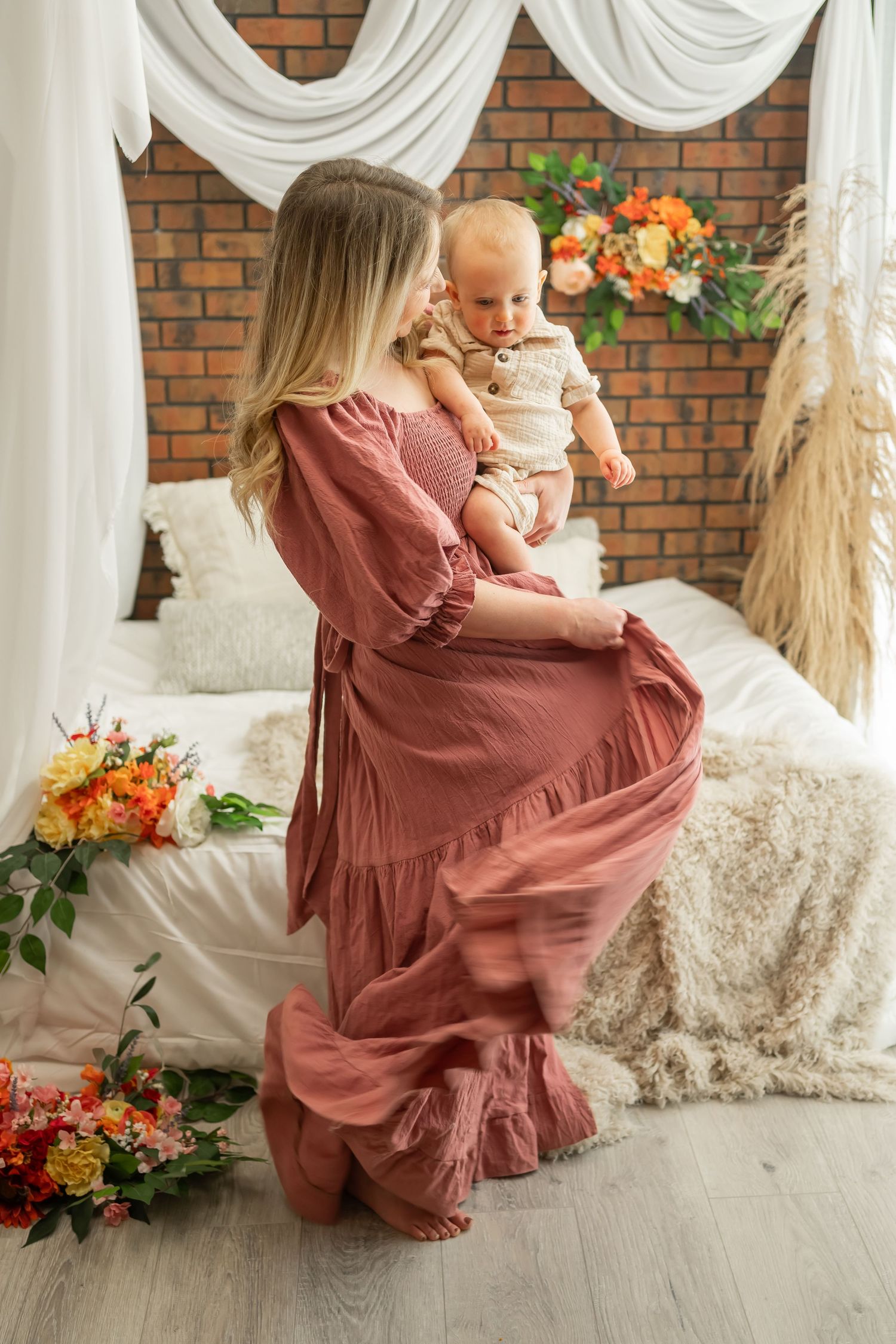 Woman in flowing mauve dress holding baby in front of rustic brick wall with floral arrangements.