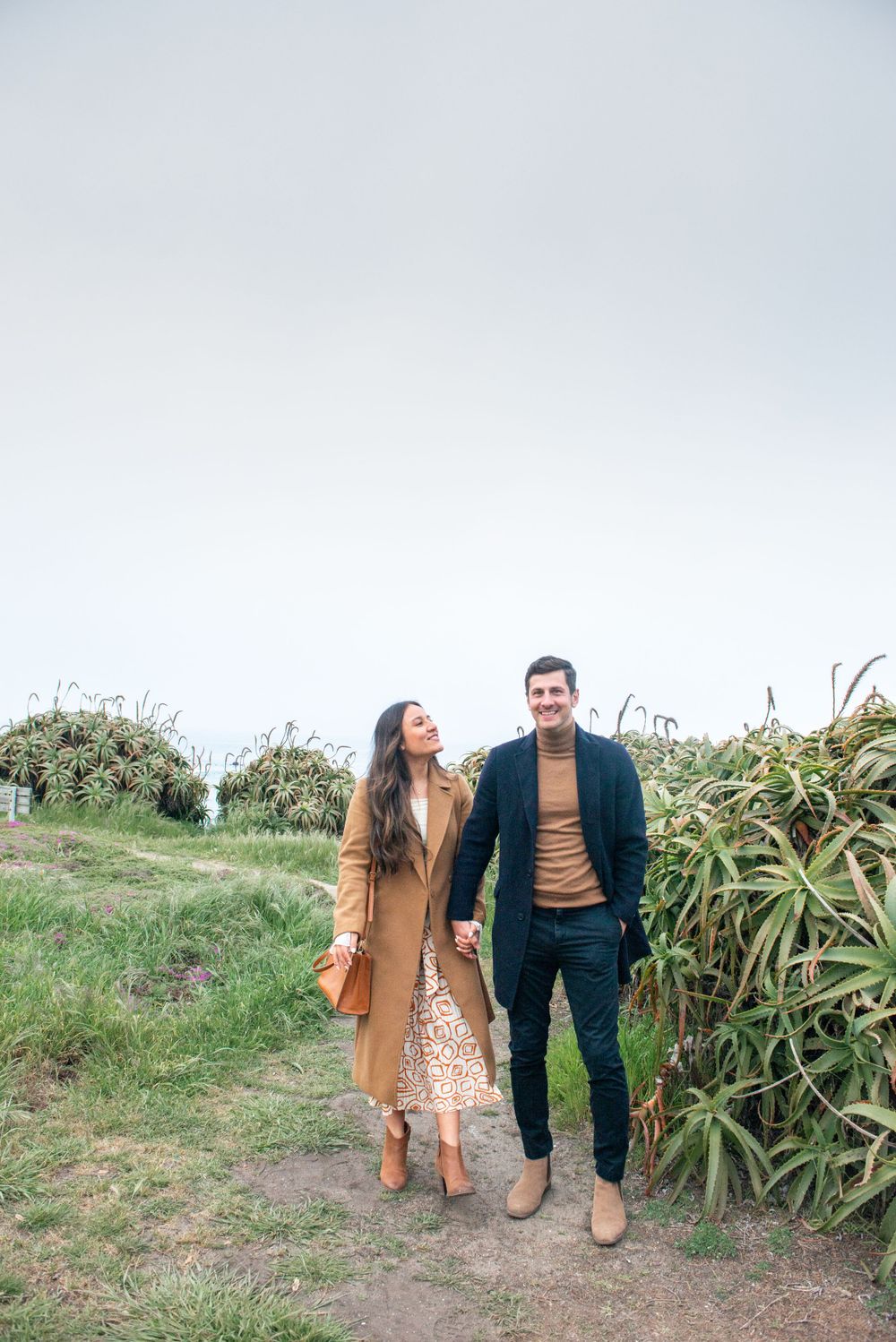 A couple in stylish autumn outfits walking together on a path with tall plants and greenery in the background.