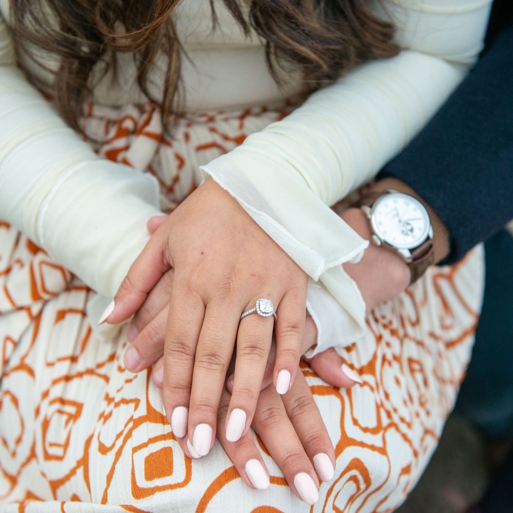 Close-up of hands displaying an engagement ring with white nail polish and a watch against an orange patterned fabric.