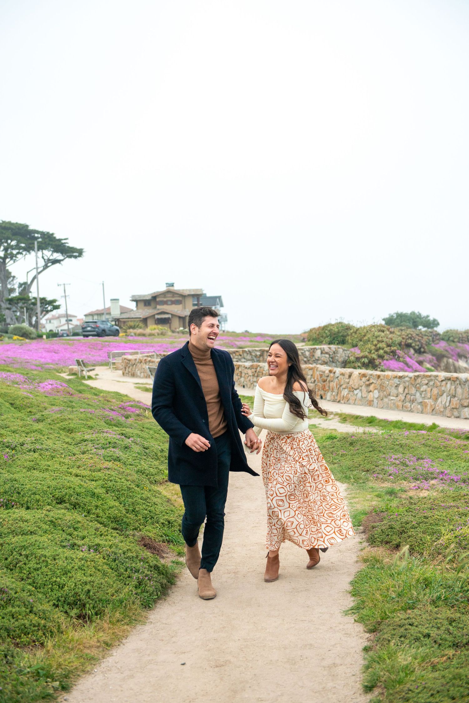 Couple walking joyfully together on a coastal path with purple wildflowers blooming along the cliffside on a bright day.