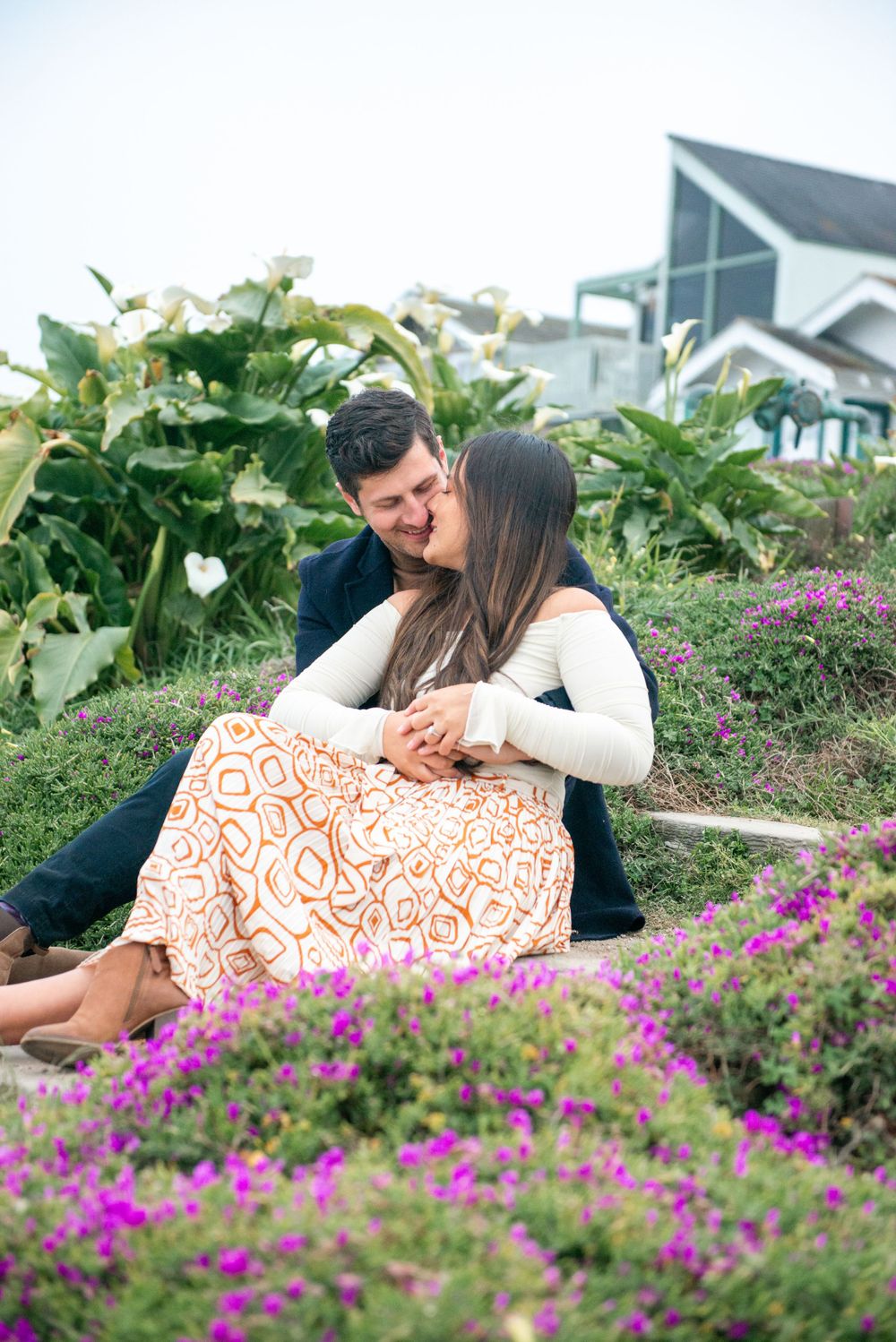 A couple embraces while sitting in a garden surrounded by vibrant purple flowers and lush greenery.