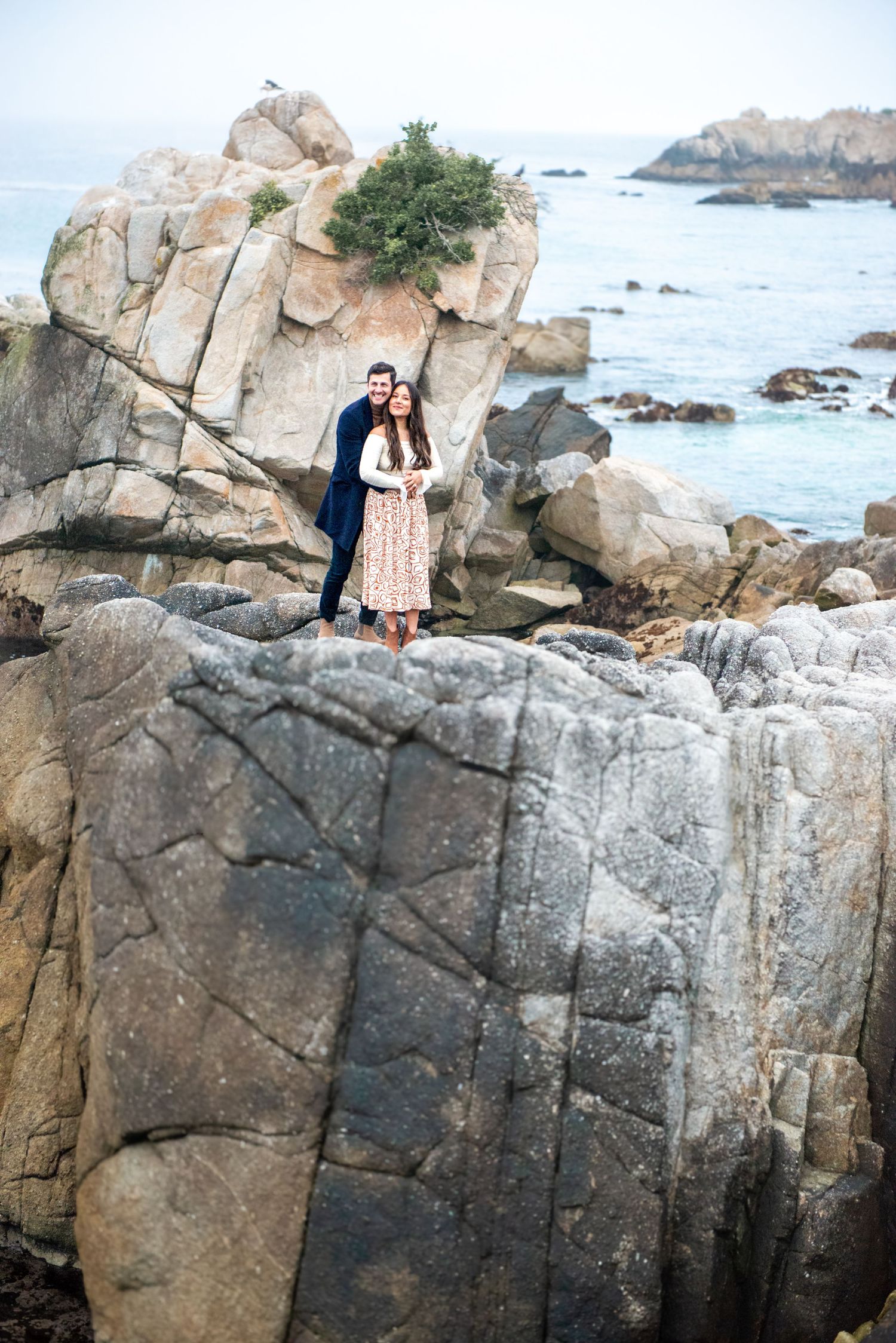 Two people stand together on dramatic coastal rocks overlooking a rocky beach and ocean view on a cloudy day.