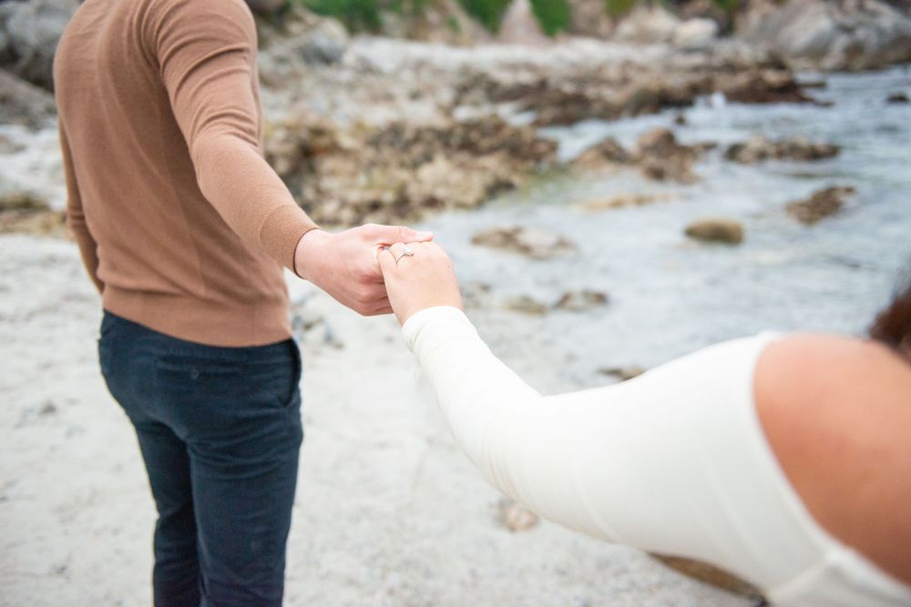 Two people clasping hands on a rocky beach with blurred background of stones and water.