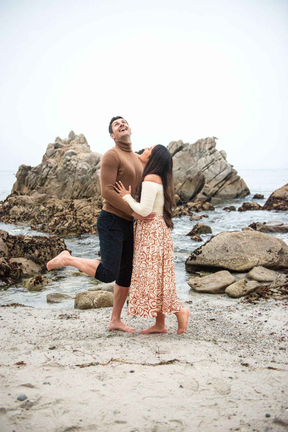 A romantic couple embraces on a rocky beach with waves crashing against coastal rocks in the background.