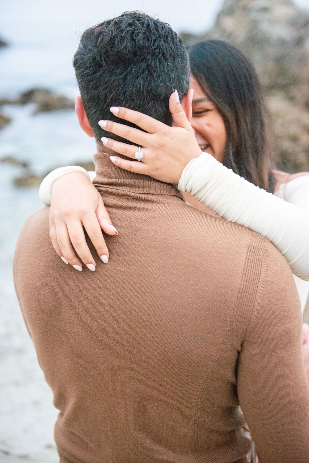 Two people embrace on a rocky beach, one wearing a brown sweater while the other wears white.