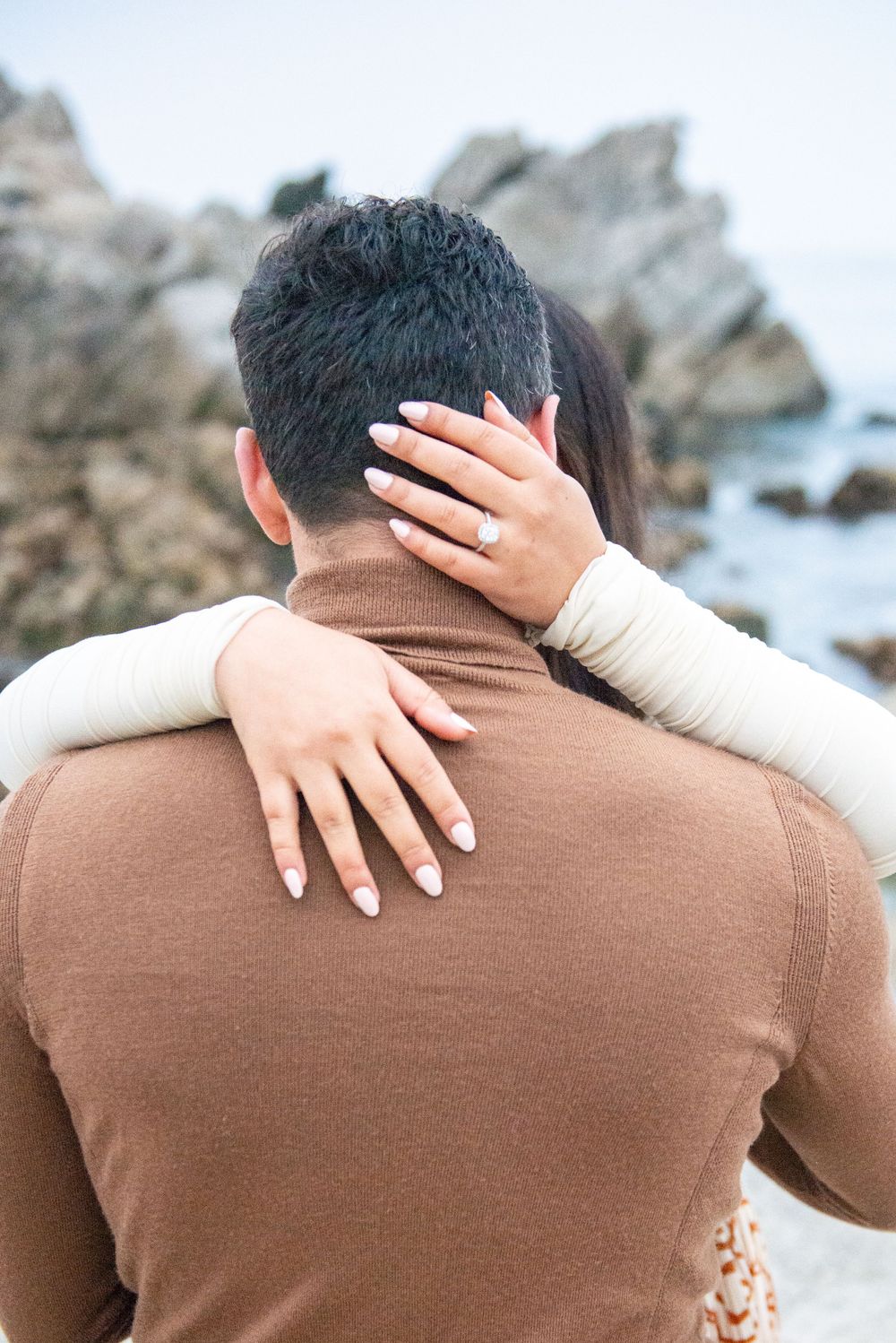 Person wearing brown sweater embraced from behind with hands visible against rocky coastal backdrop.