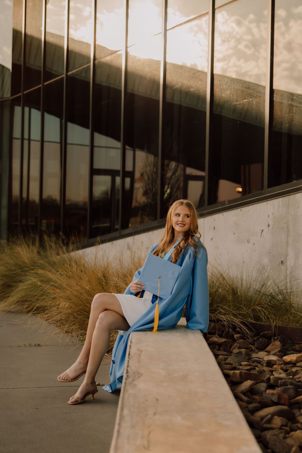A person in a light blue coat sits on a concrete ledge against a modern building with glass windows at sunset.