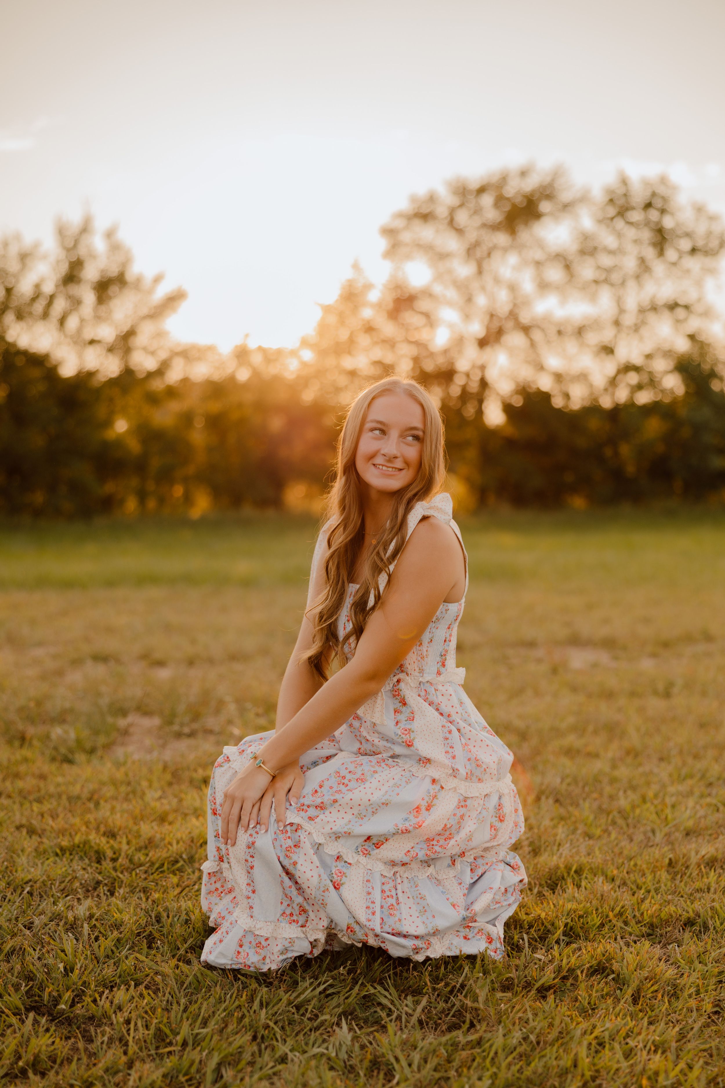 A person in a floral dress sits on grass during golden hour sunset with trees in the background.
