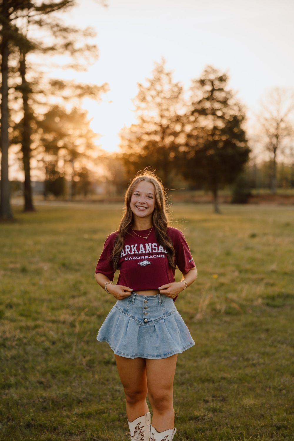 Young woman smiling in Arkansas t-shirt and denim skirt, standing in sunlit field at golden hour.