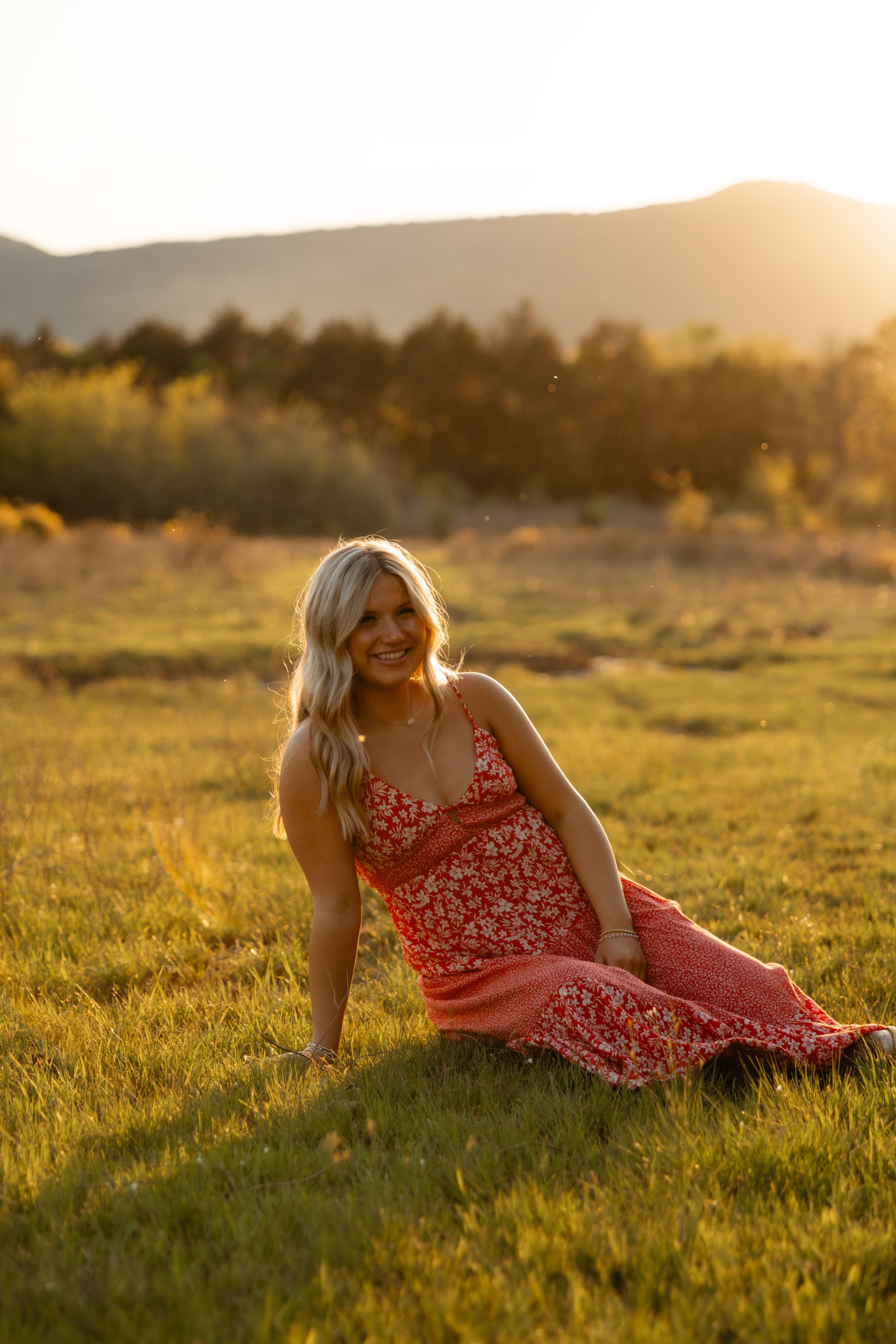 Person in red floral dress sitting in sunlit meadow with mountains in background during golden hour.