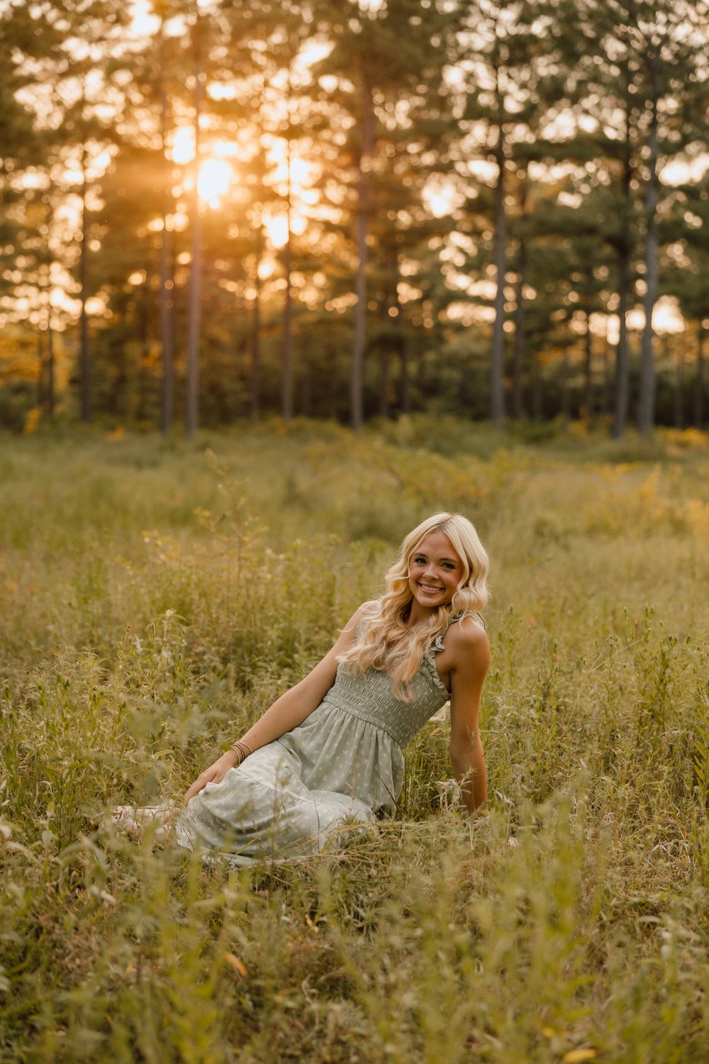 A woman in a flowy dress sits in a golden meadow at sunset with pine trees silhouetted in the background.