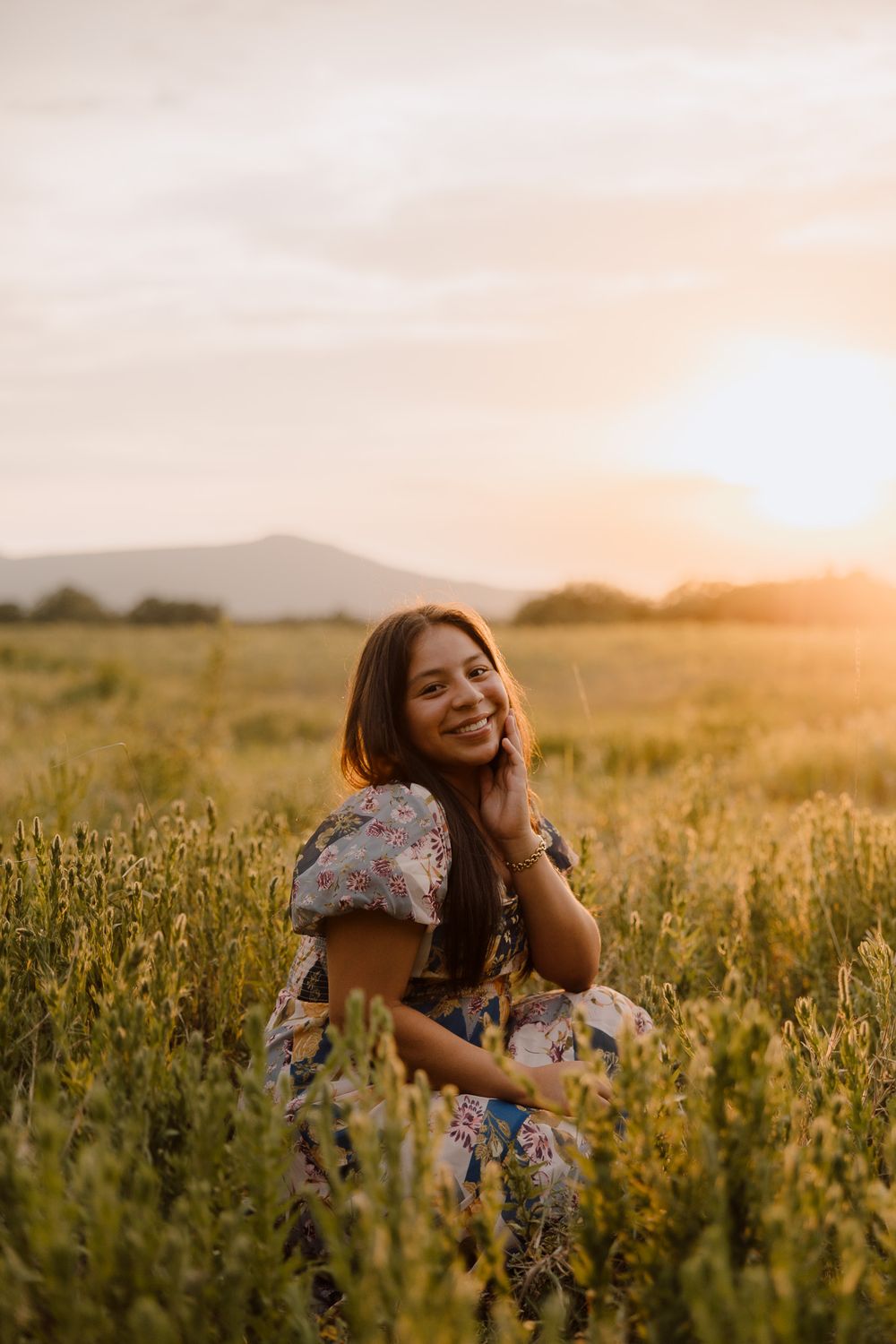 Smiling woman in floral dress sitting in golden field at sunset with mountains in background.