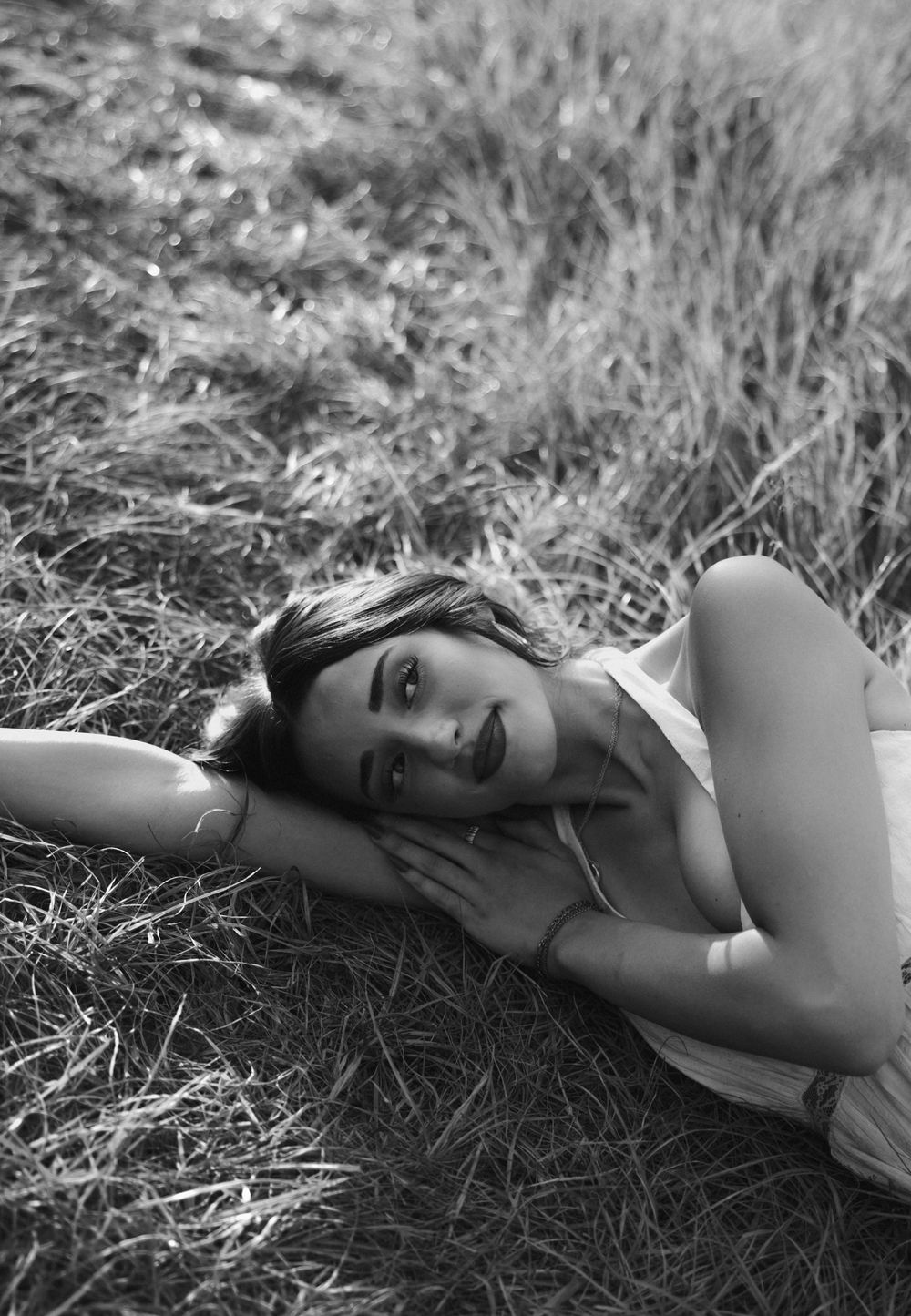 Black and white photo of a young woman lying in a grassy field, resting her face on her hand.