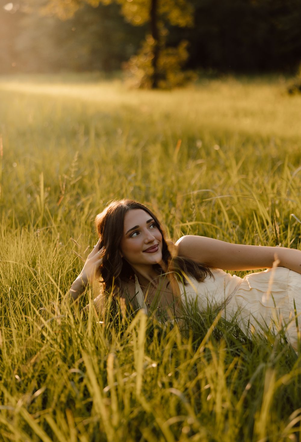 Young woman lying in golden sunlit meadow grass, smiling upward in a white dress during magic hour.