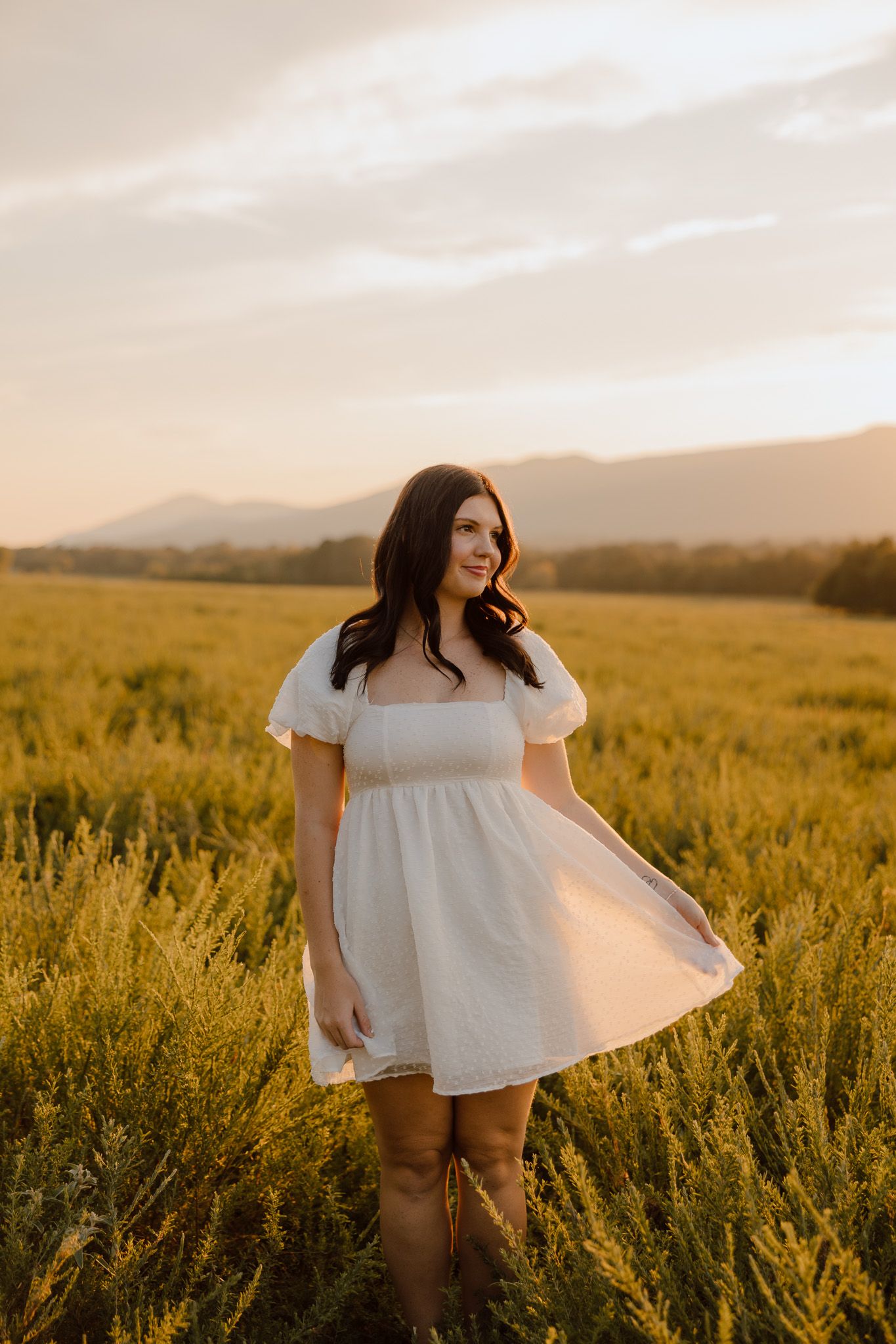 A white dress catches the golden sunset light in a field of tall grass with mountains in the background.