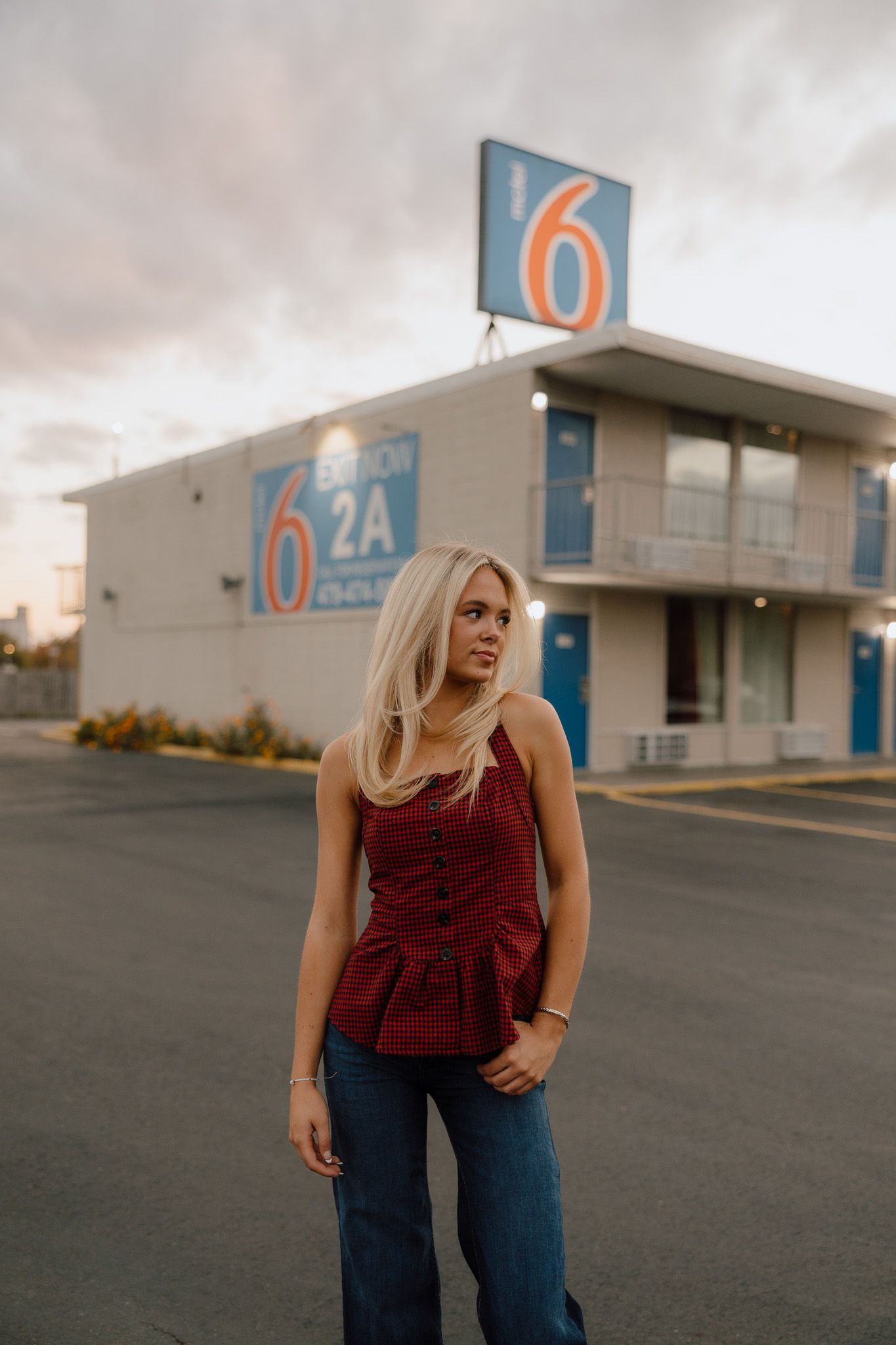 A person in a burgundy top and jeans poses in front of an industrial building with blue signage at dusk.