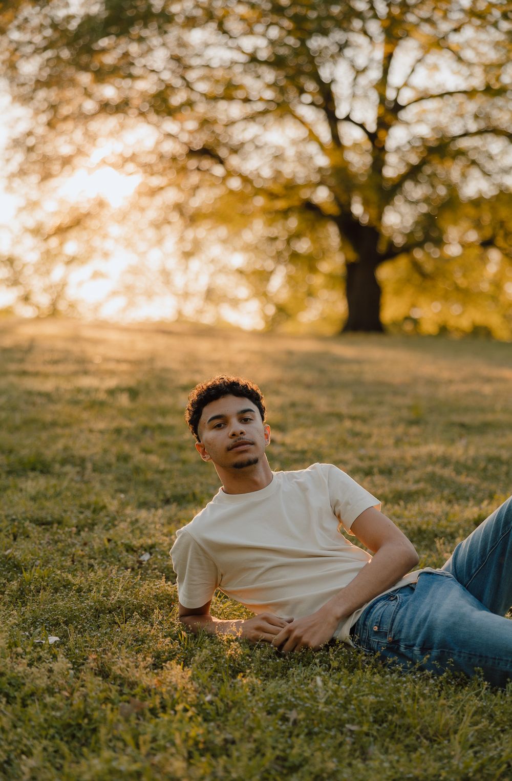 Young man in white t-shirt and jeans relaxing on green grass at golden hour with a large tree in the background.