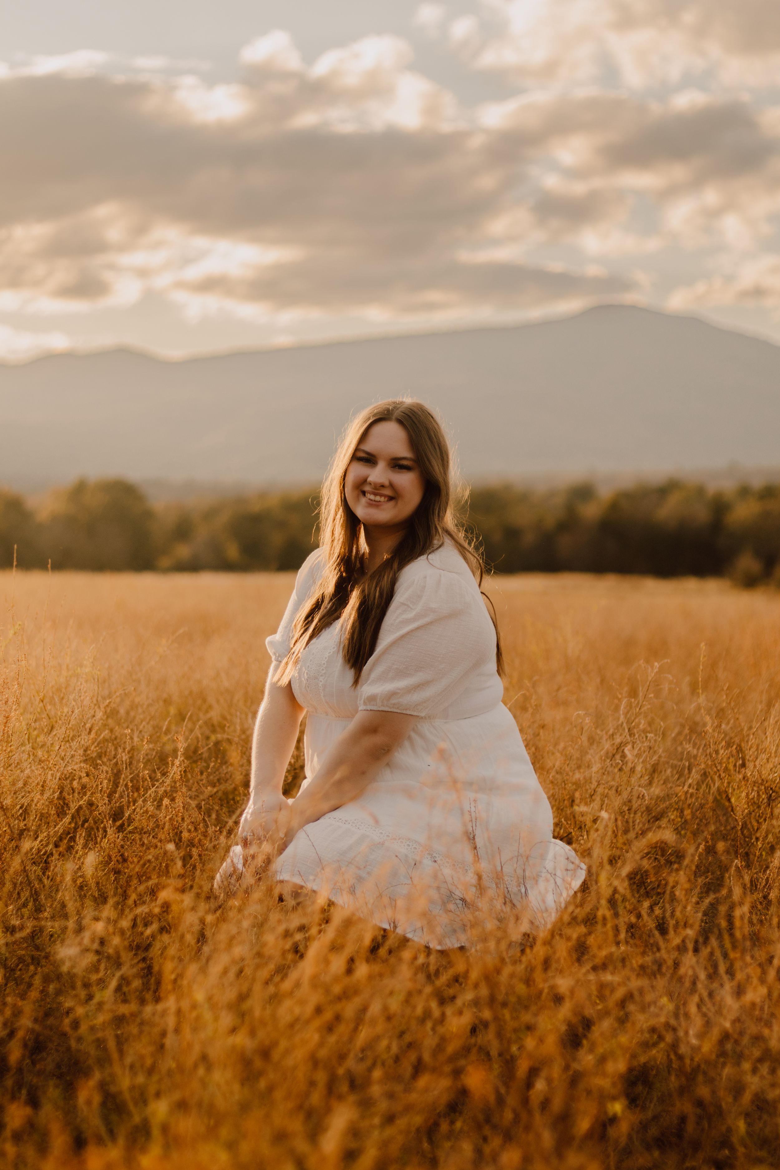 Person in white dress sitting in golden wheat field during sunset with mountains in background.