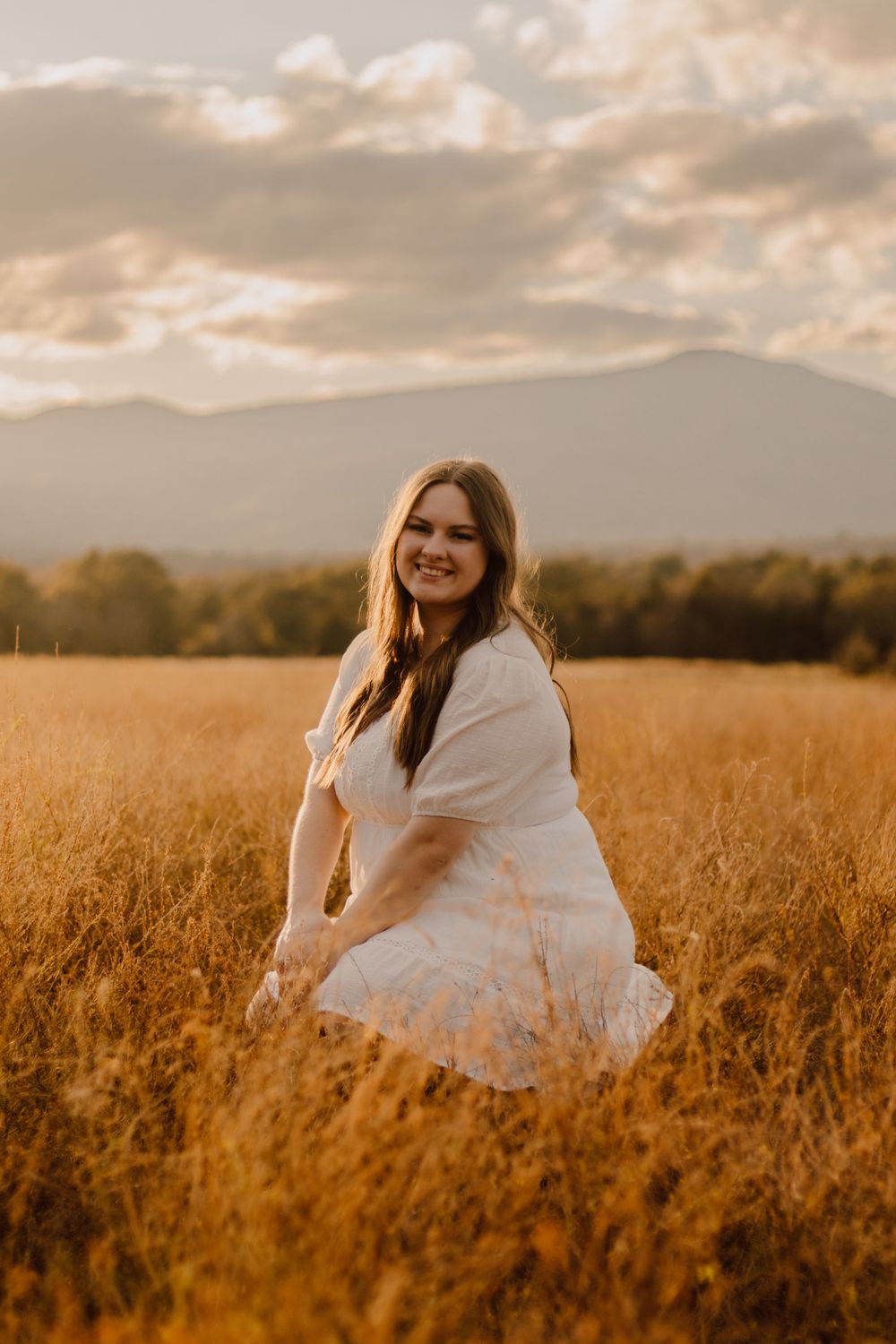 Person in white dress sitting in golden wheat field during sunset with mountains in background.