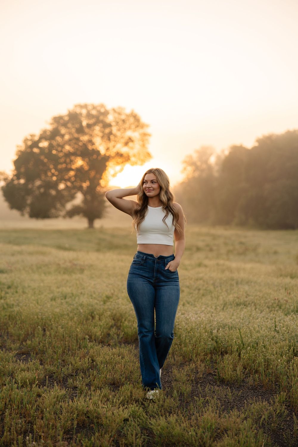 A woman in jeans and white crop top poses in a grassy field at sunset with golden light and trees in background.