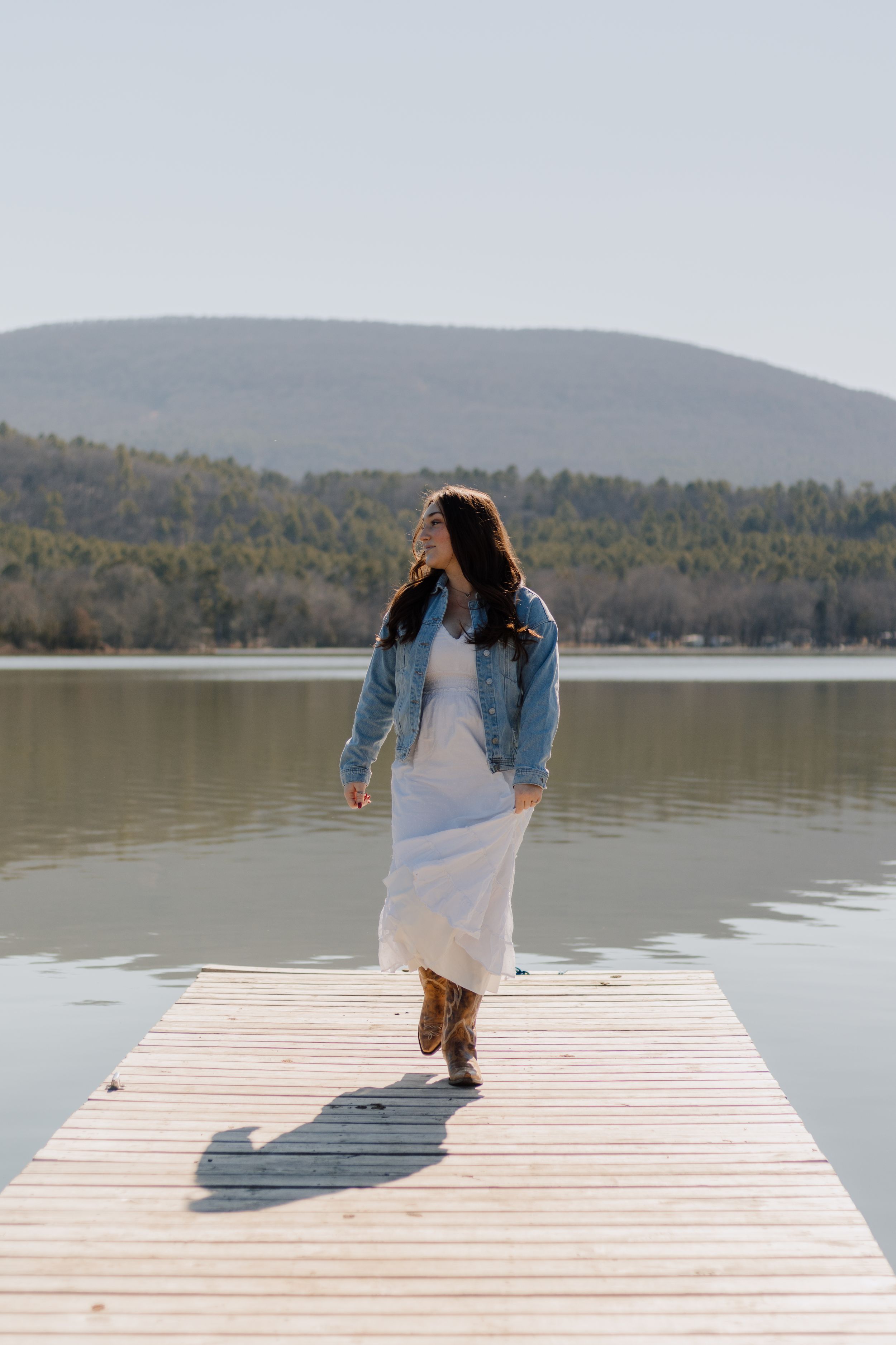 Person in white dress and denim jacket walking on wooden dock overlooking mountain lake.