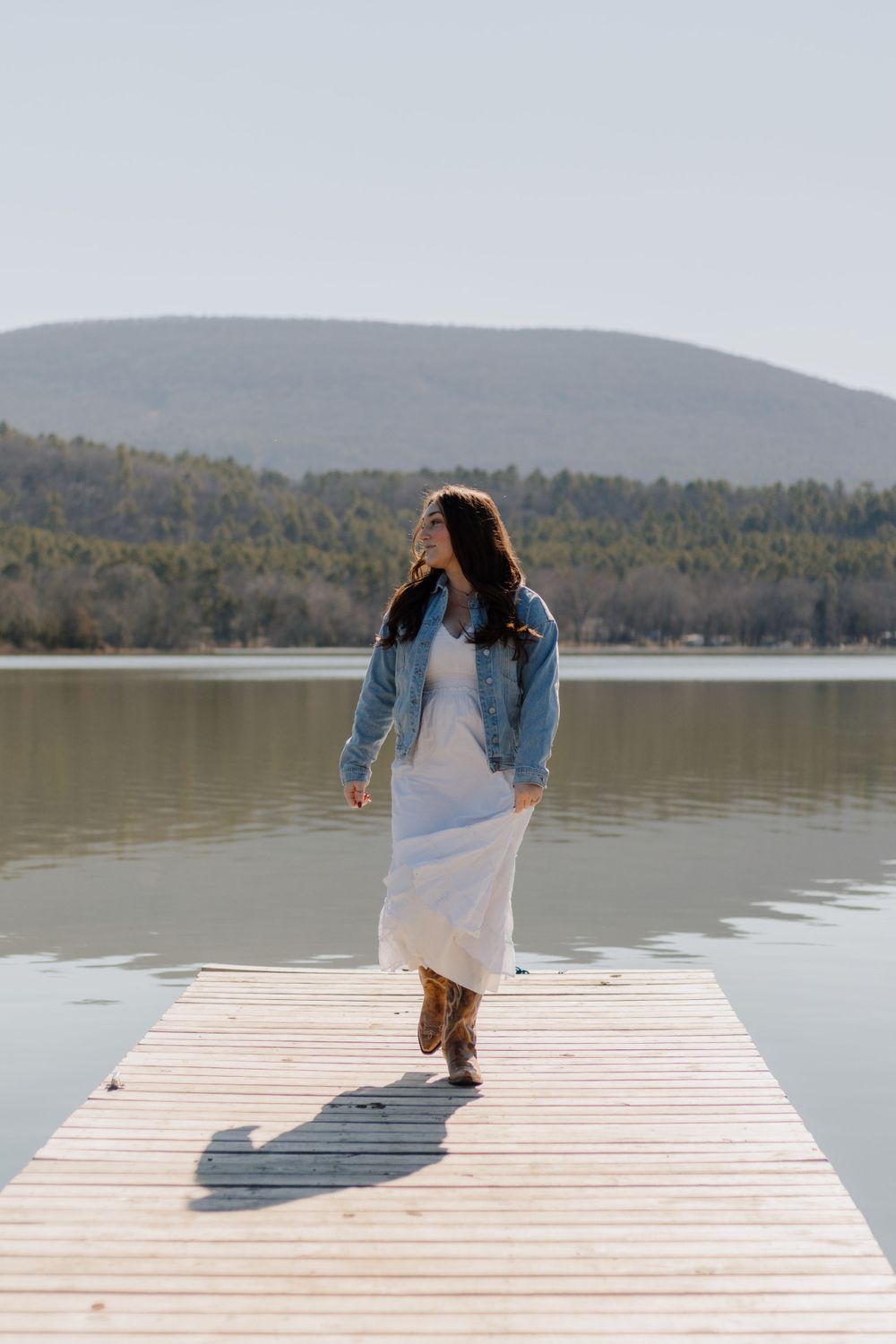 Person in white dress and denim jacket walking on wooden dock overlooking mountain lake.