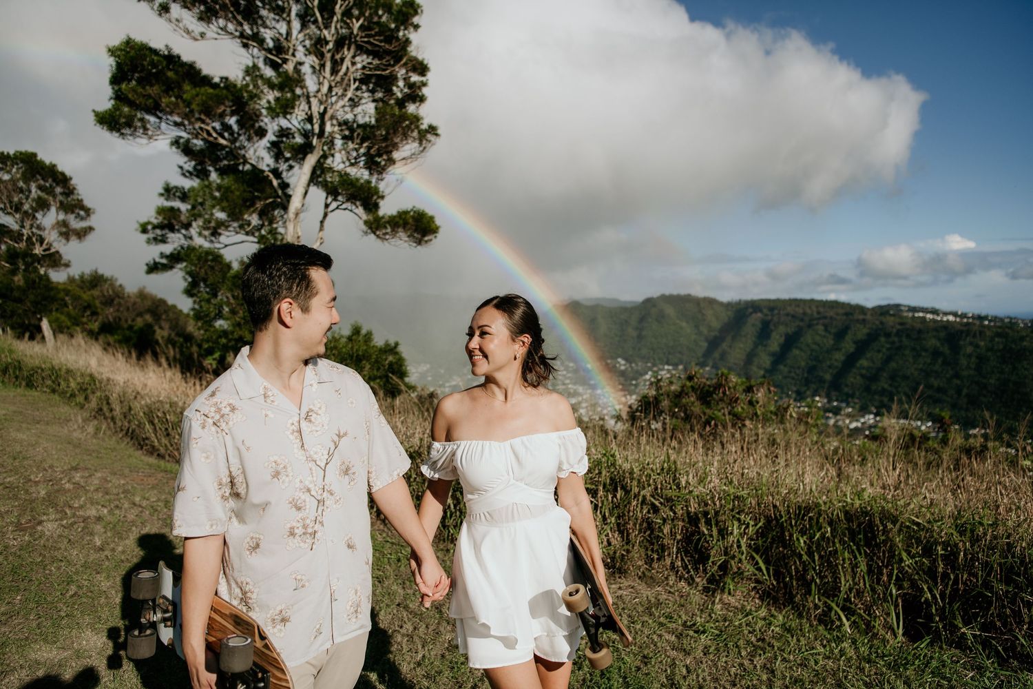 A couple in wedding attire walks hand in hand along a scenic coastal road during golden hour.