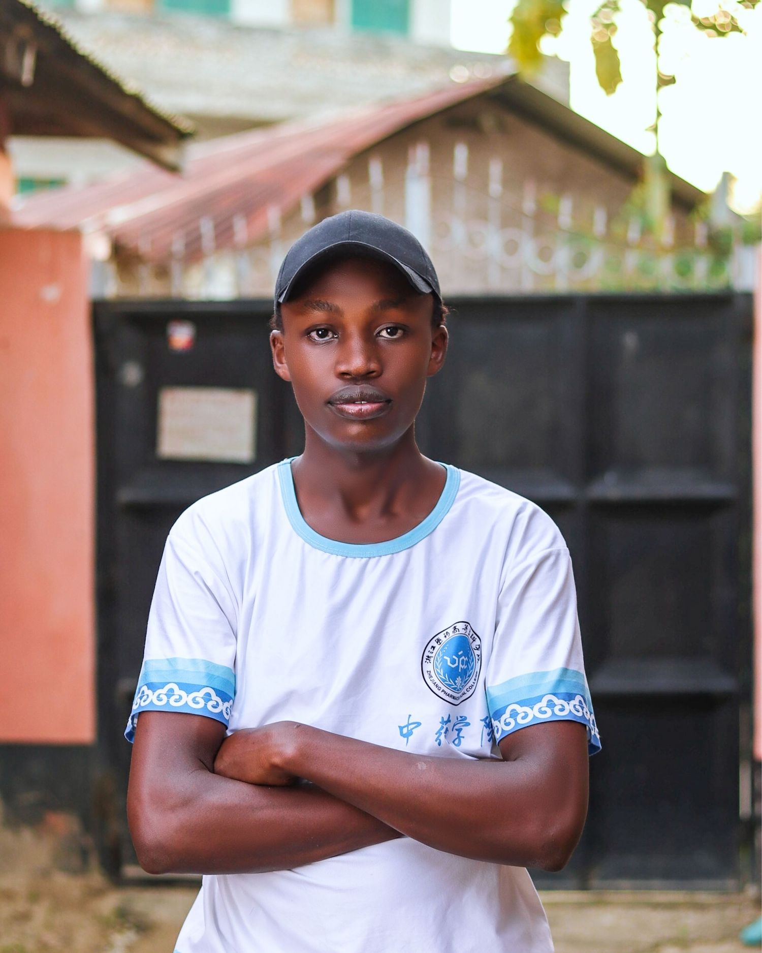 A young man poses with arms folded, behind him blurred out gate that is black in colour