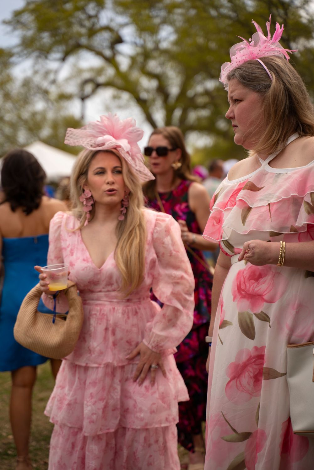 Women in pink floral dresses and fascinators chat while holding drinks at an outdoor social event.
