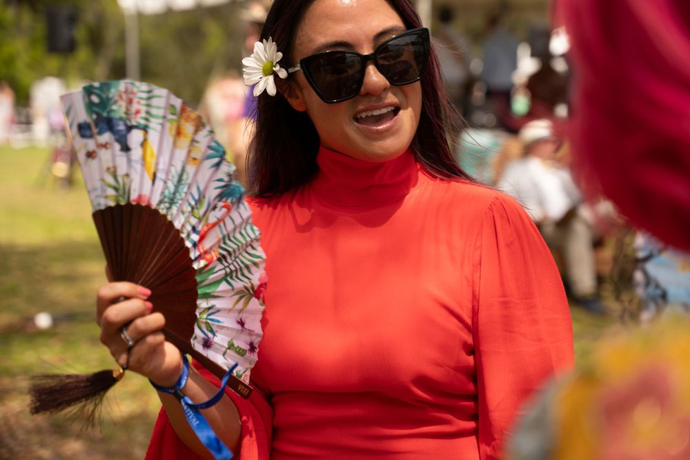 A woman in a bright red top holds a decorative folding fan at an outdoor event.