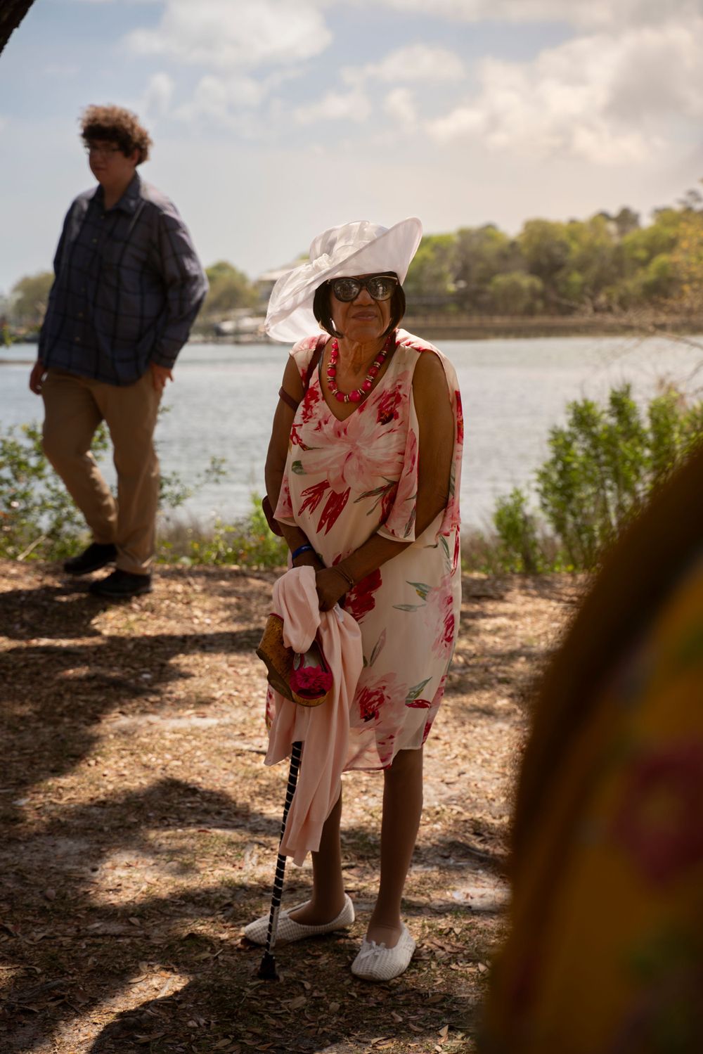 A elderly black lady in a floral dress and white hat stands near a waterfront using a walking cane at Airlie Luncheon Garden Party.