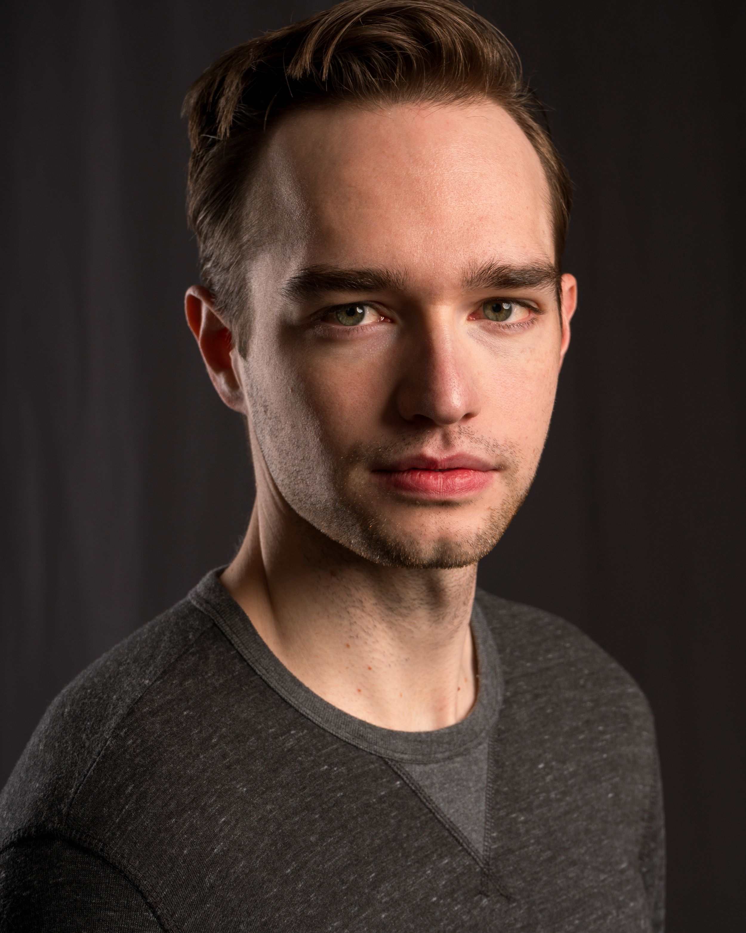 Professional headshot portrait of a young person in a gray sweater against a dark background.