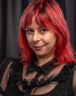 Studio shot of someone with bright red hair wearing a black lace top against a dark background.