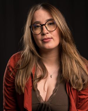 Professional portrait of someone in glasses and a red jacket against a dark background.